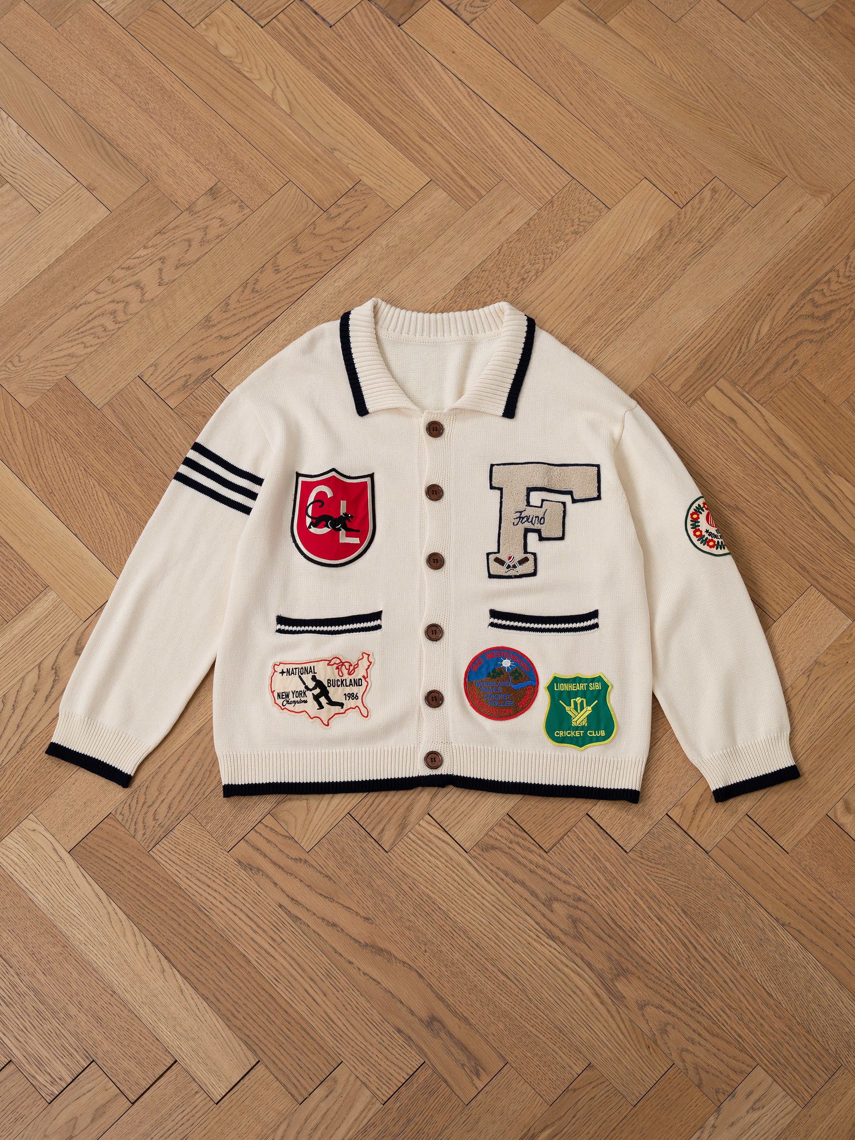 The York Varsity Patch Collared Cardigan by FOUND evokes collegiate nostalgia with black trim, buttons, and patches like a red shield, large "F," and circular badges displayed on a wooden herringbone floor.