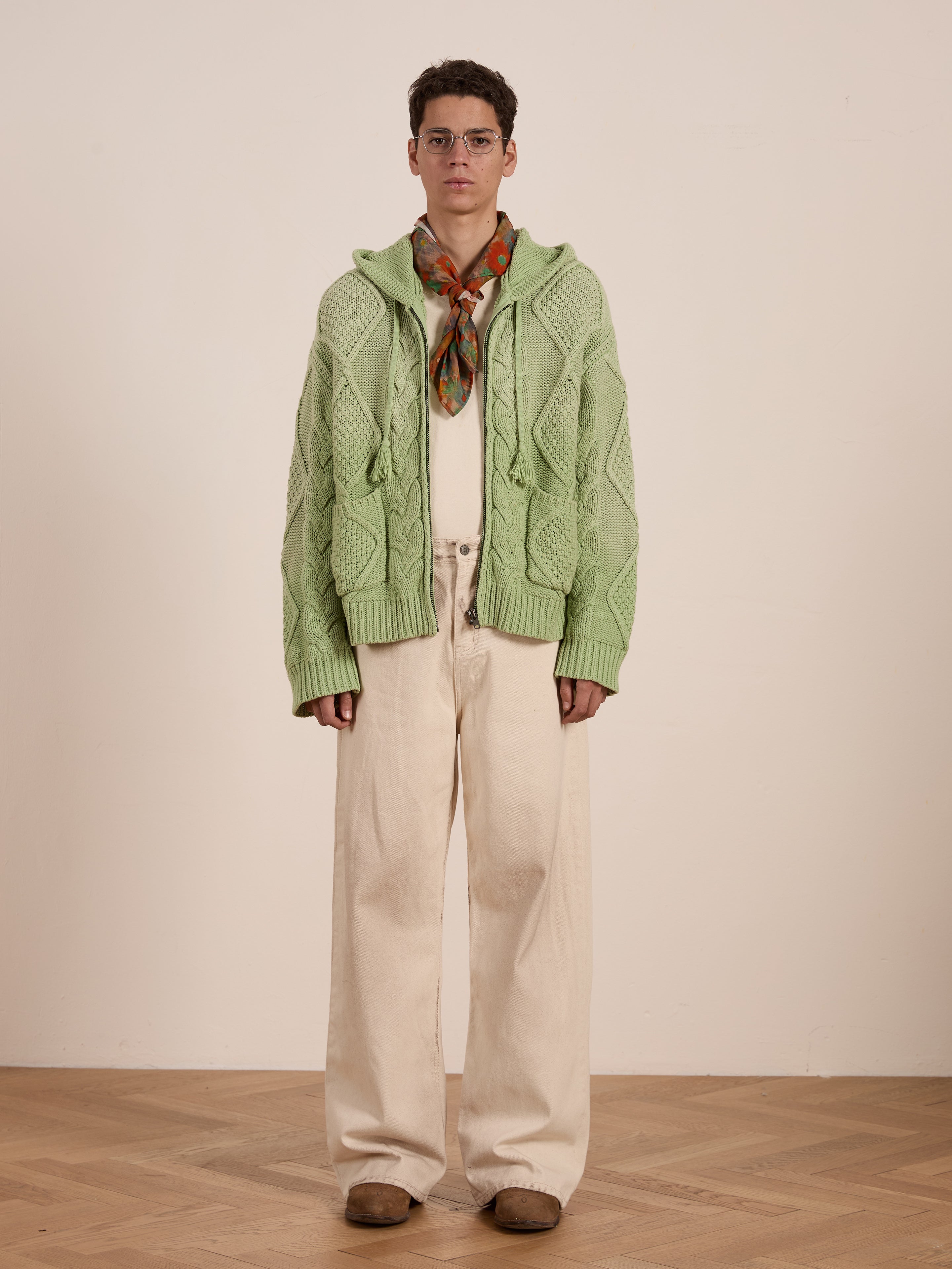 A person dons glasses and a light green Found Zip Up Cable Knit Hoodie over a wooden floor. With beige pants and a colorful scarf, the unisex relaxed fit embodies casual elegance against the plain backdrop.