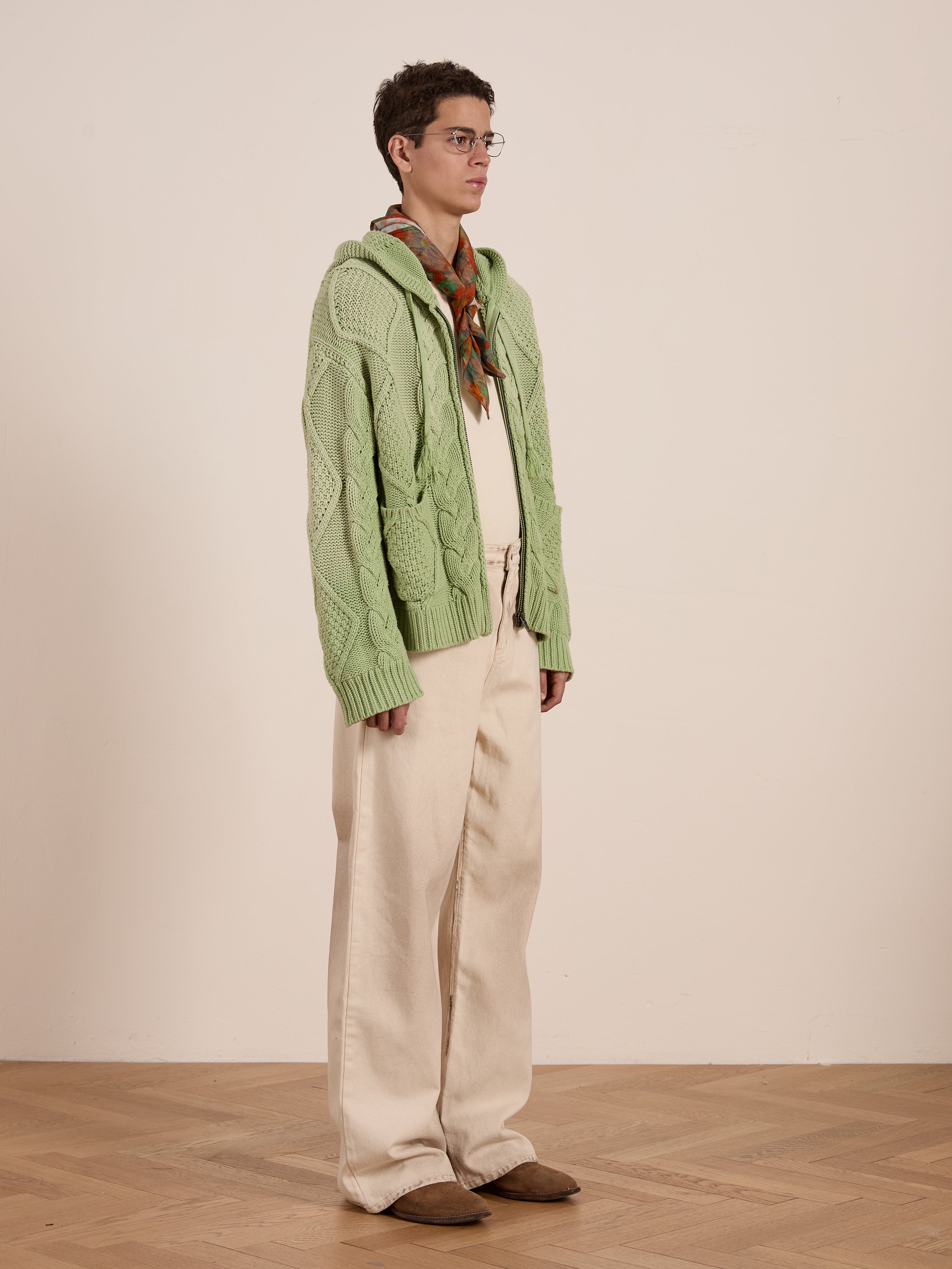 Indoors, a person wears a FOUND light green Zip Up Cable Knit Hoodie paired with cream pants and a patterned scarf, exuding an effortlessly cozy unisex relaxed fit.