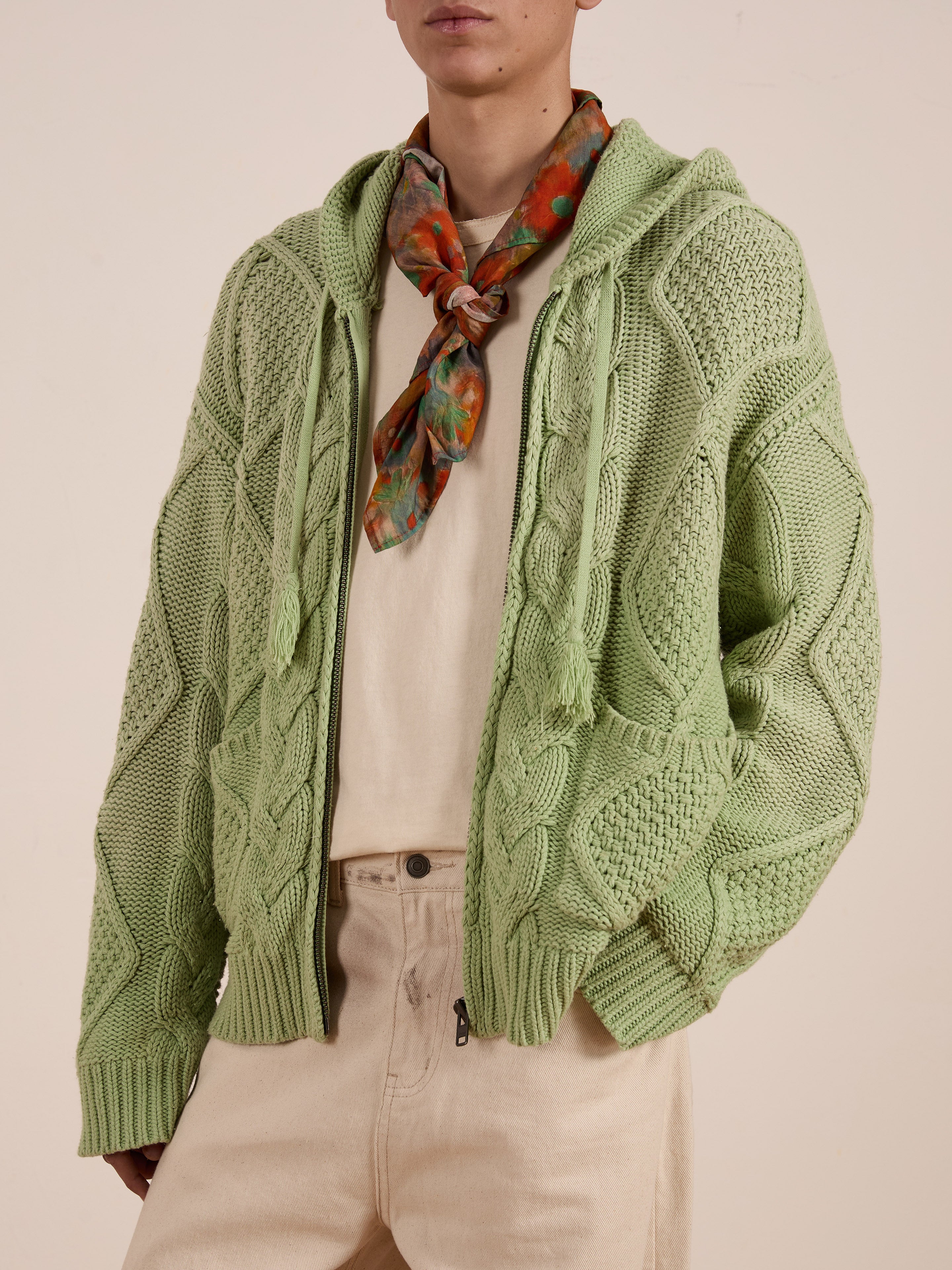 A person is wearing a green FOUND Zip Up Cable Knit Hoodie in a unisex relaxed fit, paired with a beige shirt, beige pants, and a multicolored neck scarf.