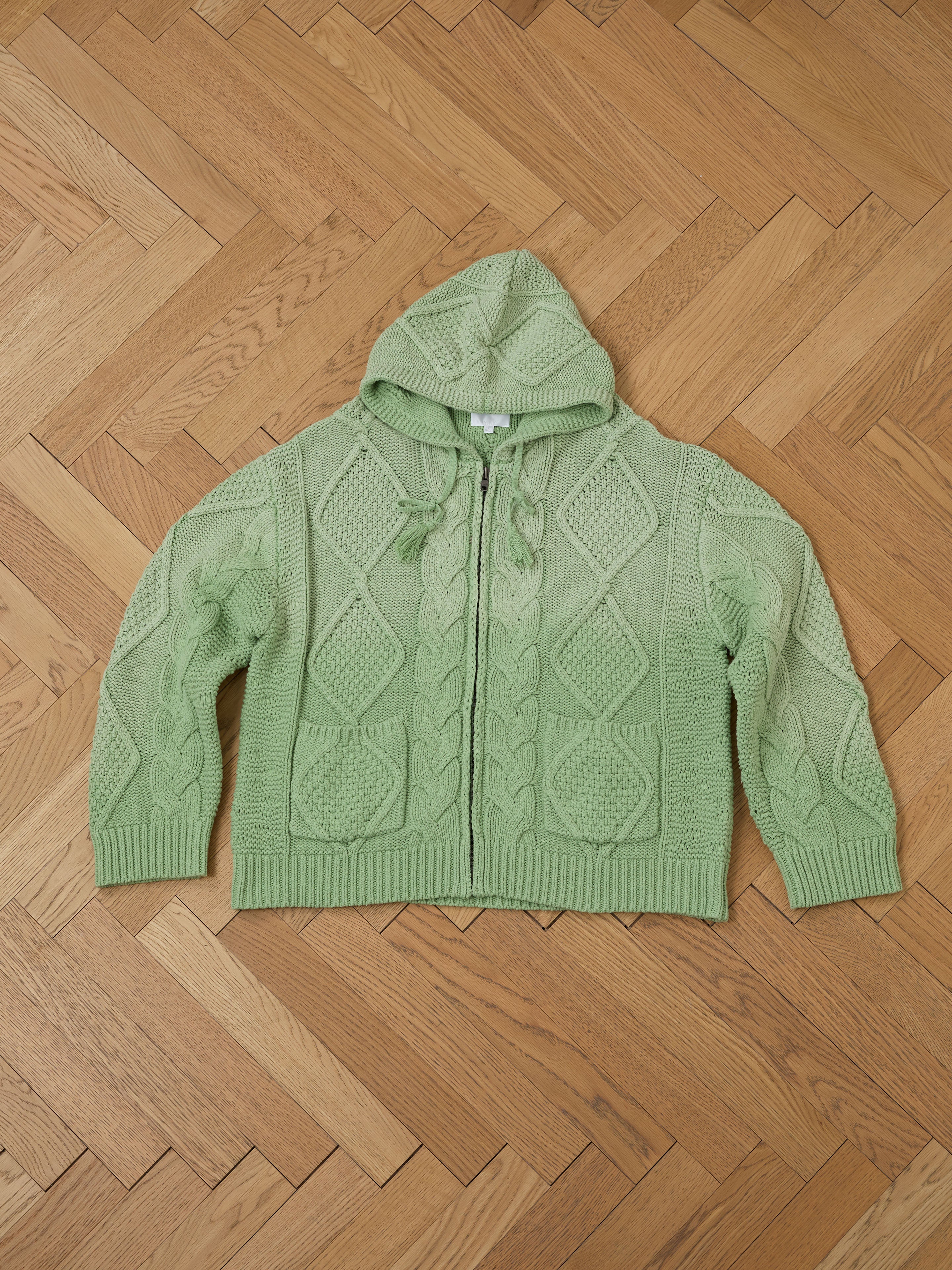 Found's Zip Up Cable Knit Hoodie, in a green cotton blend with a unisex relaxed fit, is laid flat on a wooden herringbone floor.