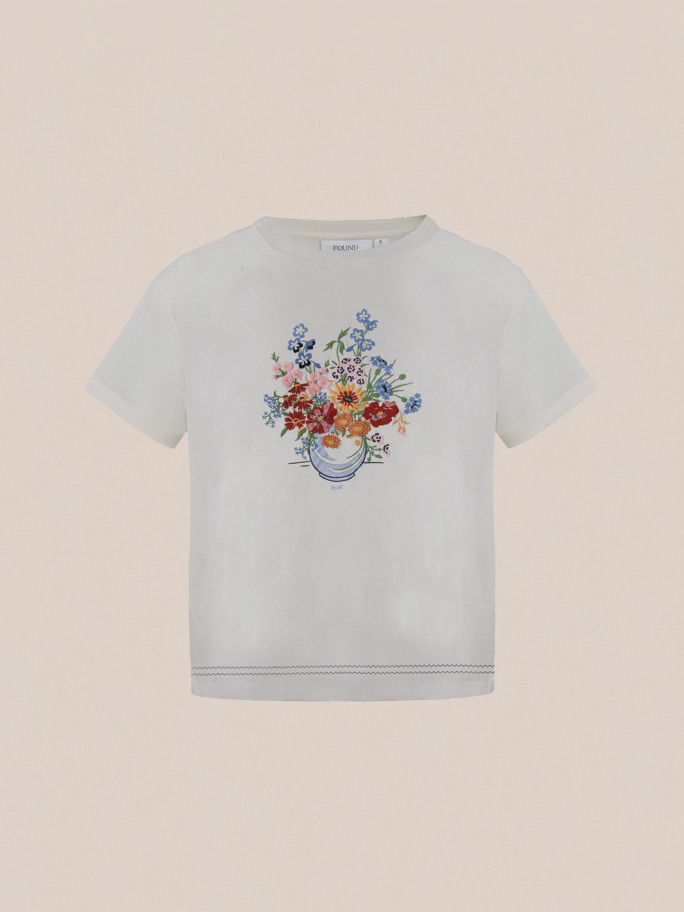 The "Bouquet Flowers Tee" by Found is a white T-shirt featuring a vibrant graphic of pink, red, and blue flowers arranged in a vase at the center. It also includes decorative stitching near the hem that is reminiscent of traditional Punjab Pakistan Phulkari embroidery, set against a light background.