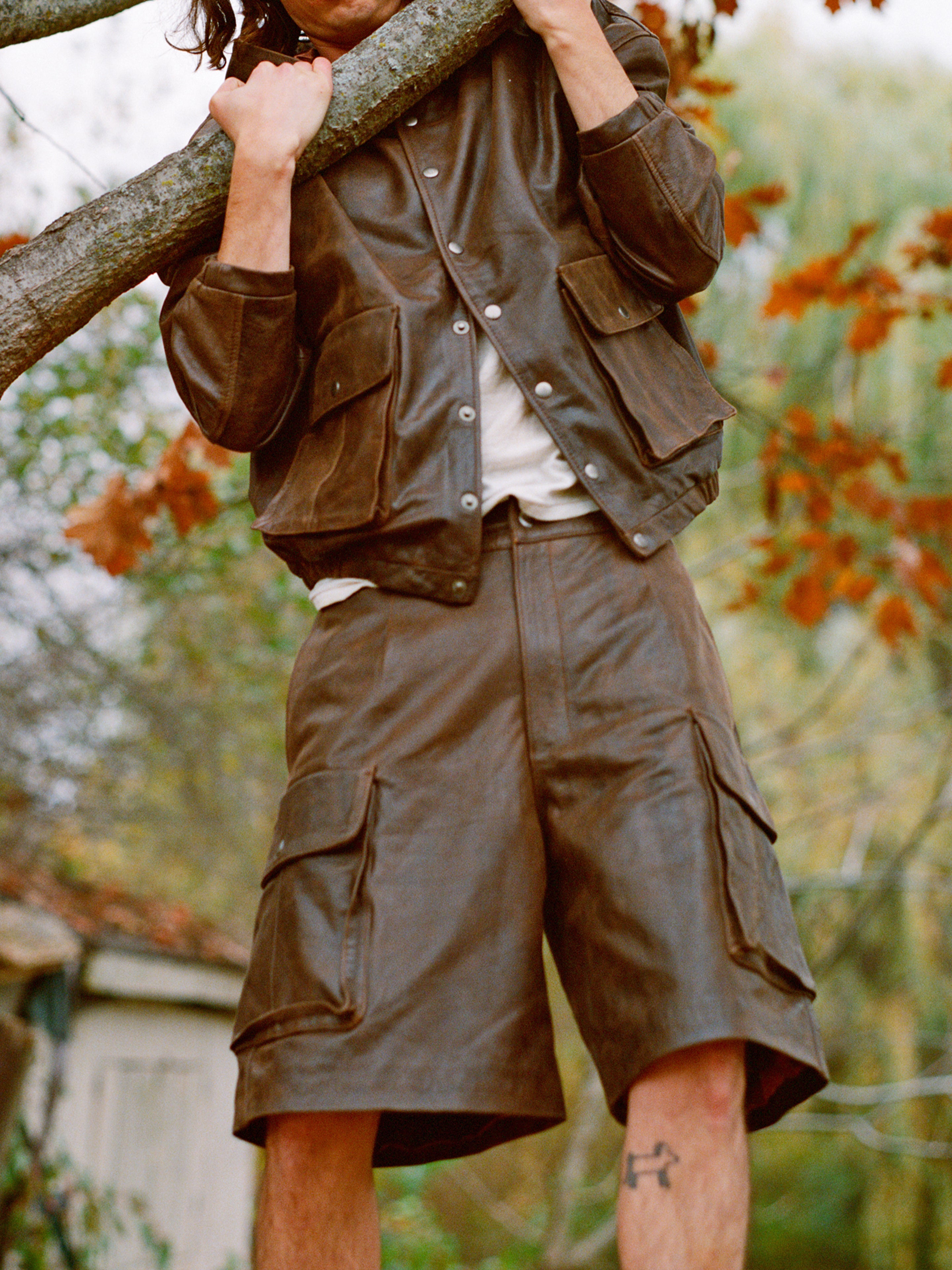 Wearing FOUND's Dusk Distressed Genuine Leather Cargo Shorts, a person climbs a tree branch amidst autumn foliage, highlighting the utilitarian roots of their style.