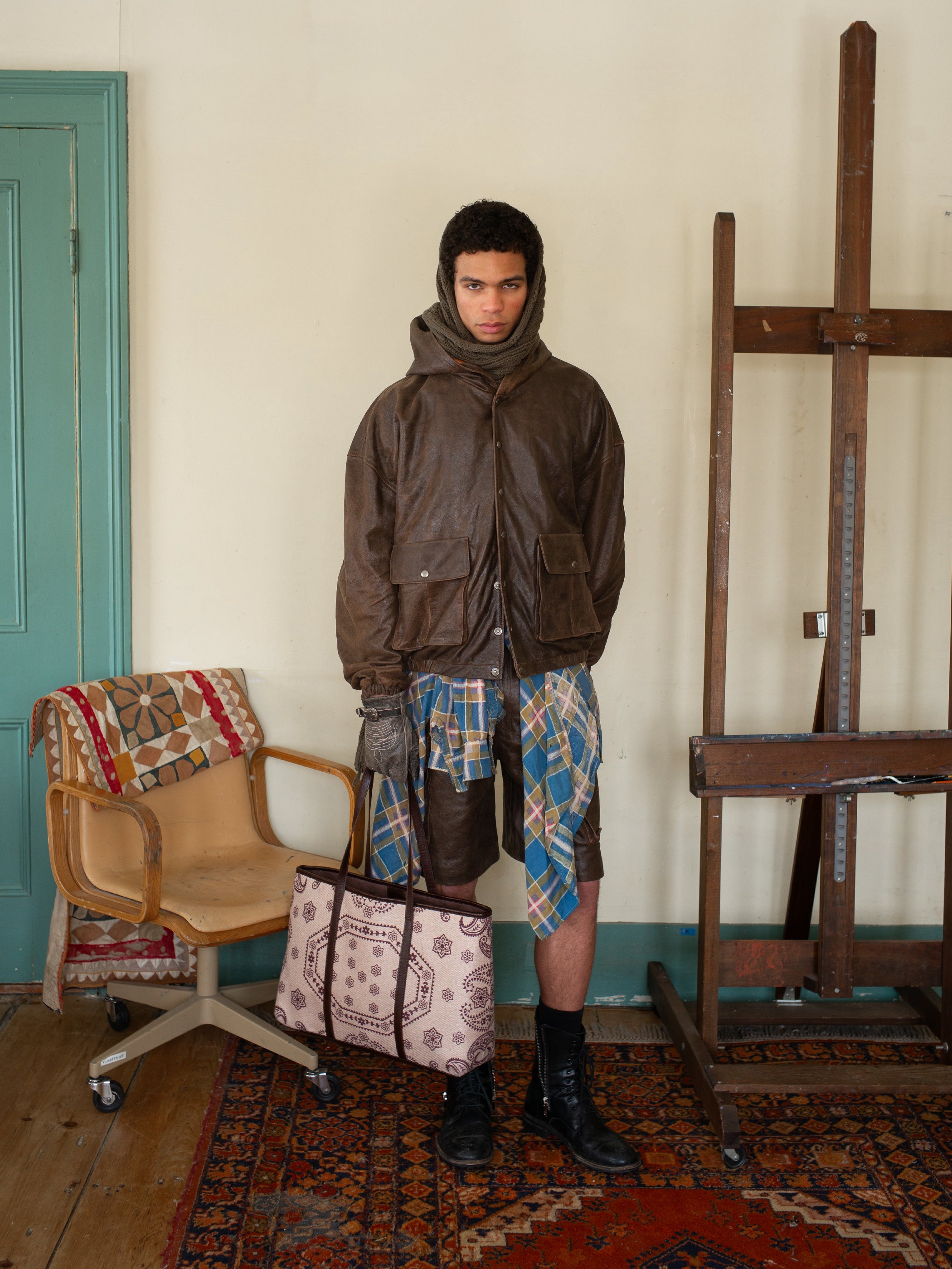 A person wearing a FOUND Dusk Distressed Genuine Leather Hoodie and plaid shorts stands indoors, holding a patterned bag next to a chair and easel.