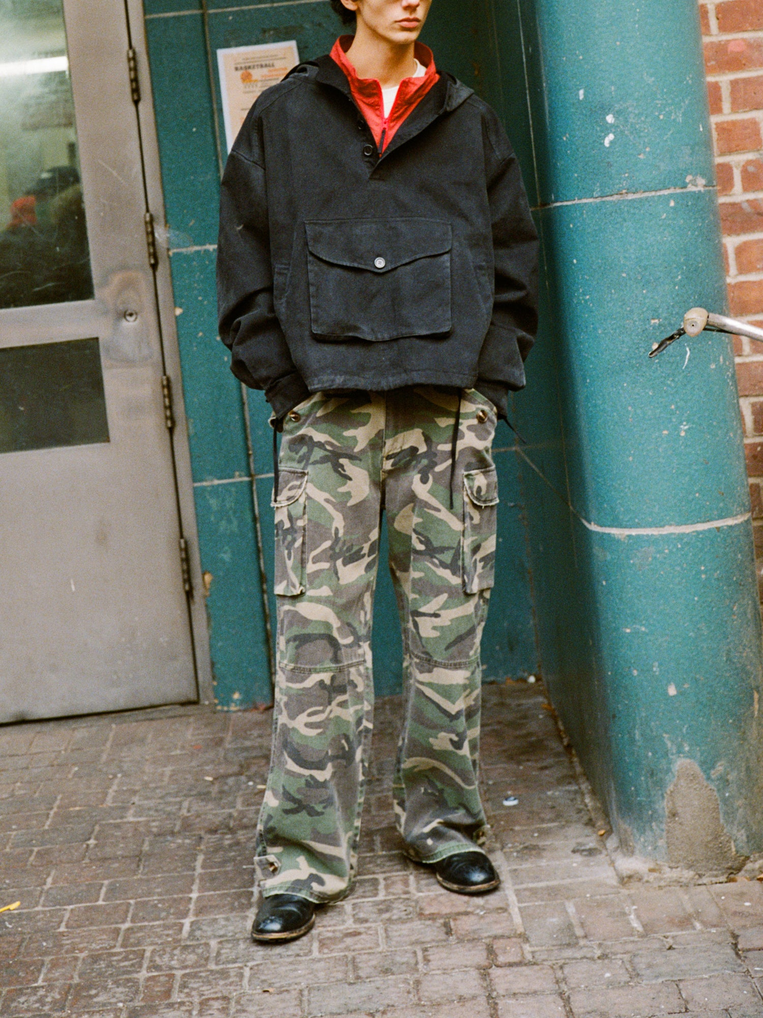 Wearing FOUND's Camouflage Twill Utility Cargo Pants with a black jacket and red shirt, a person stands near a green pillar and brick wall, adding black shoes for a vintage look.