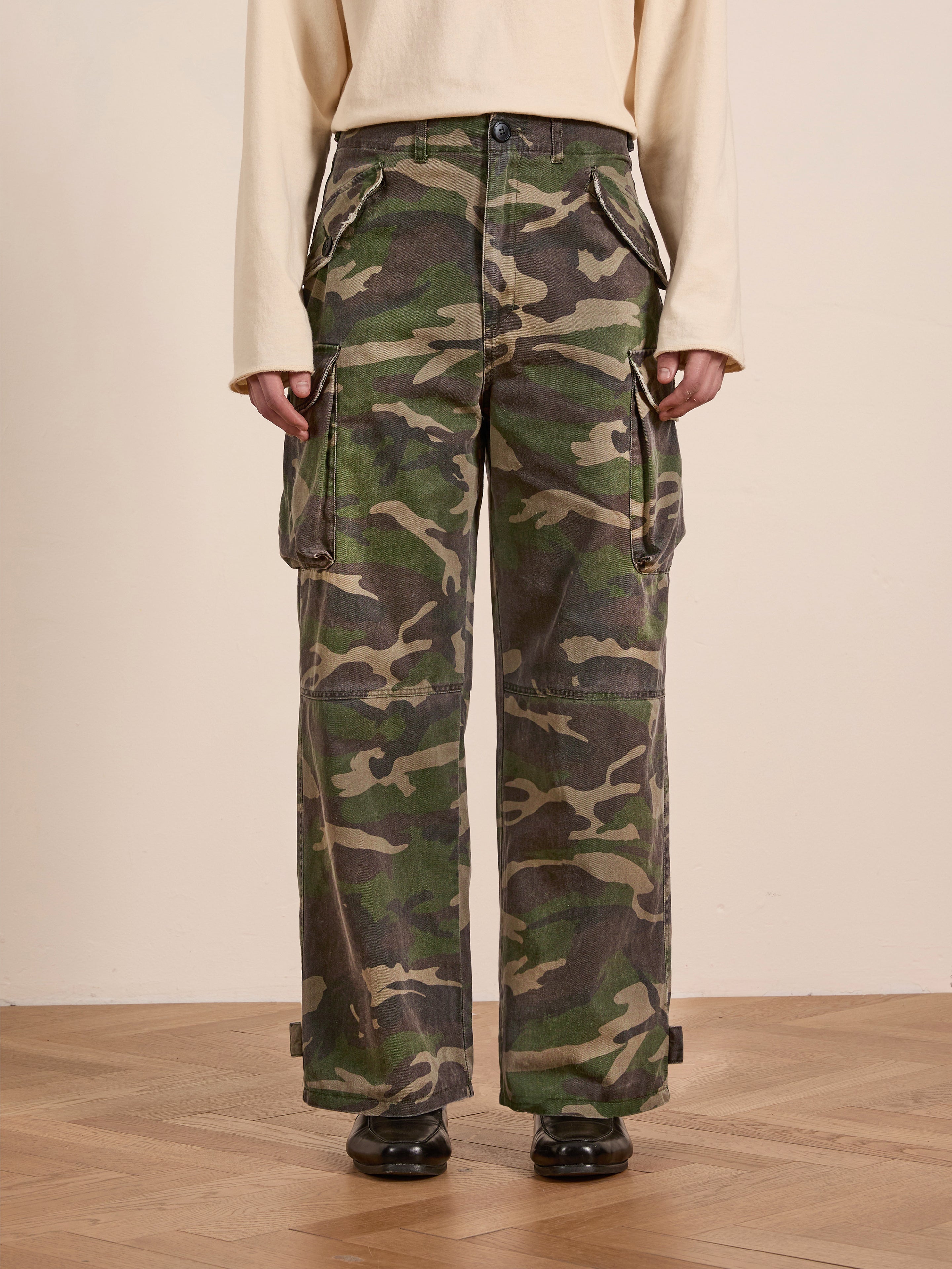 A person stands on a wooden floor, wearing FOUND's Camouflage Twill Utility Cargo Pants with a beige long-sleeve top and black shoes, creating an effortlessly stylish vintage look.