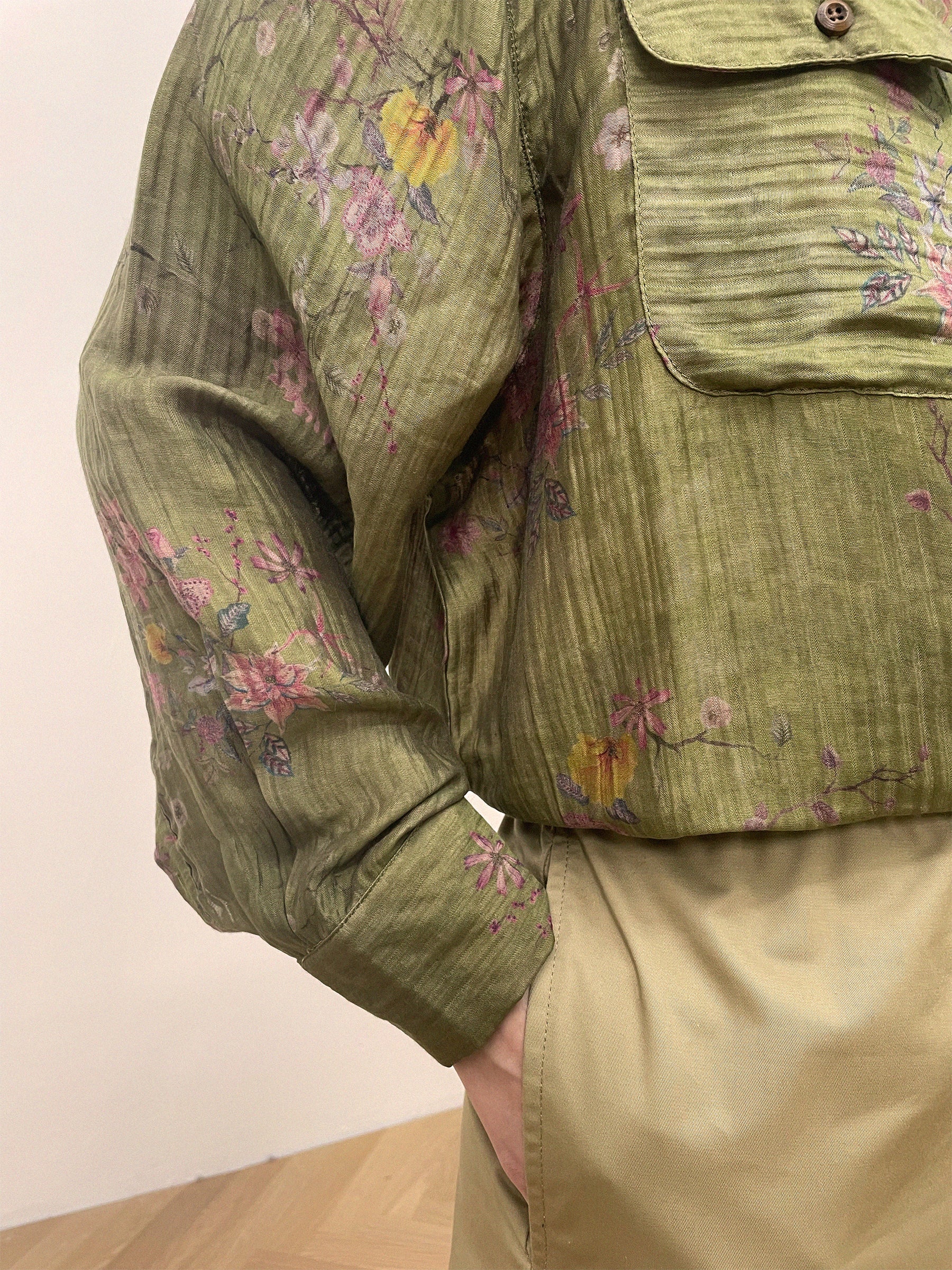 Close-up of a person wearing a green Floral Moss LS Camp Shirt by Found with dual chest pockets and beige pants, one hand casually tucked in their pocket, standing on a wooden floor.