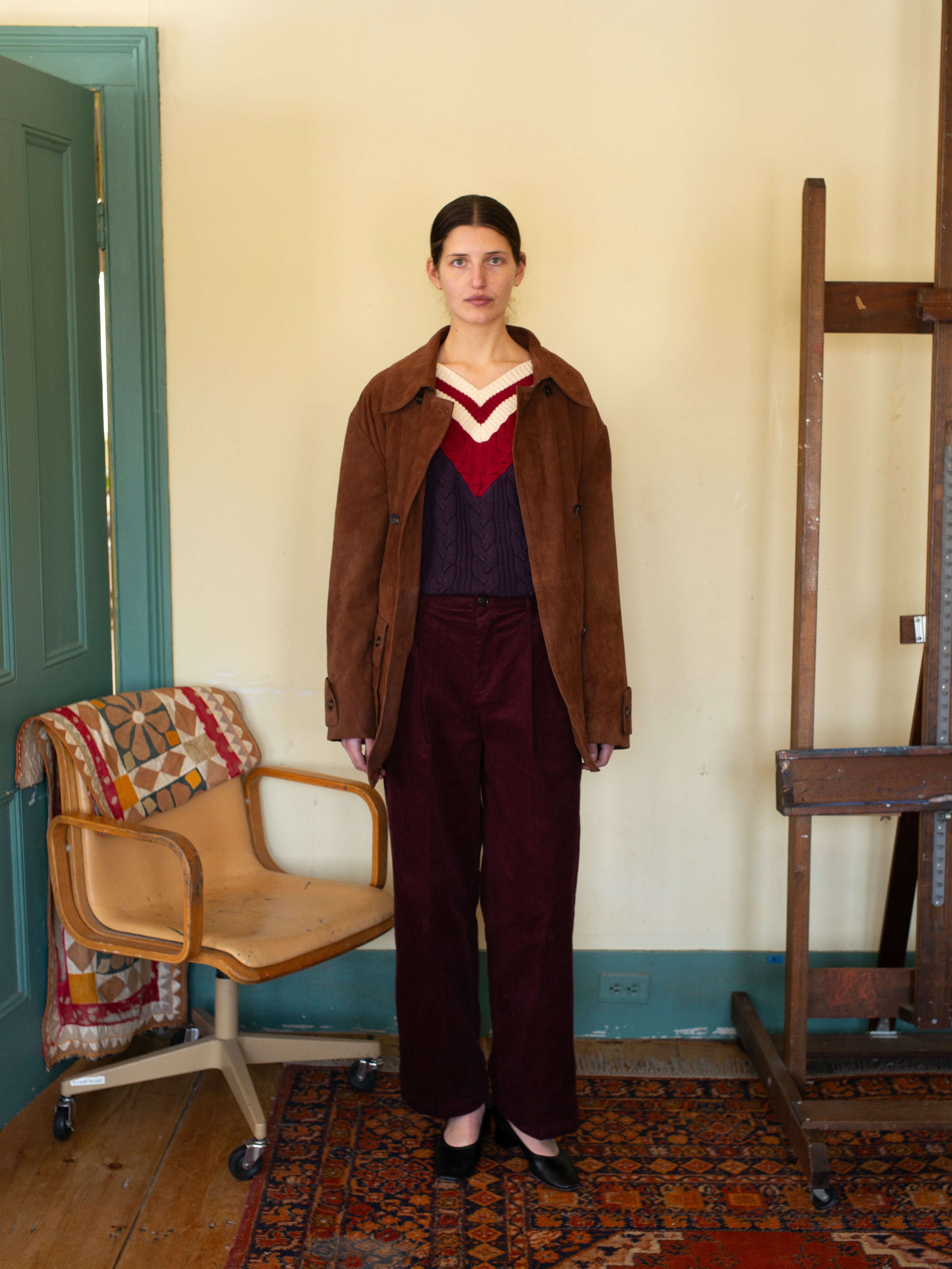 Indoors, a V-Neck Cricket Sweater from FOUND is worn with dark pants beside a wooden easel and a chair draped with a cotton quilt. The floor features a patterned rug.