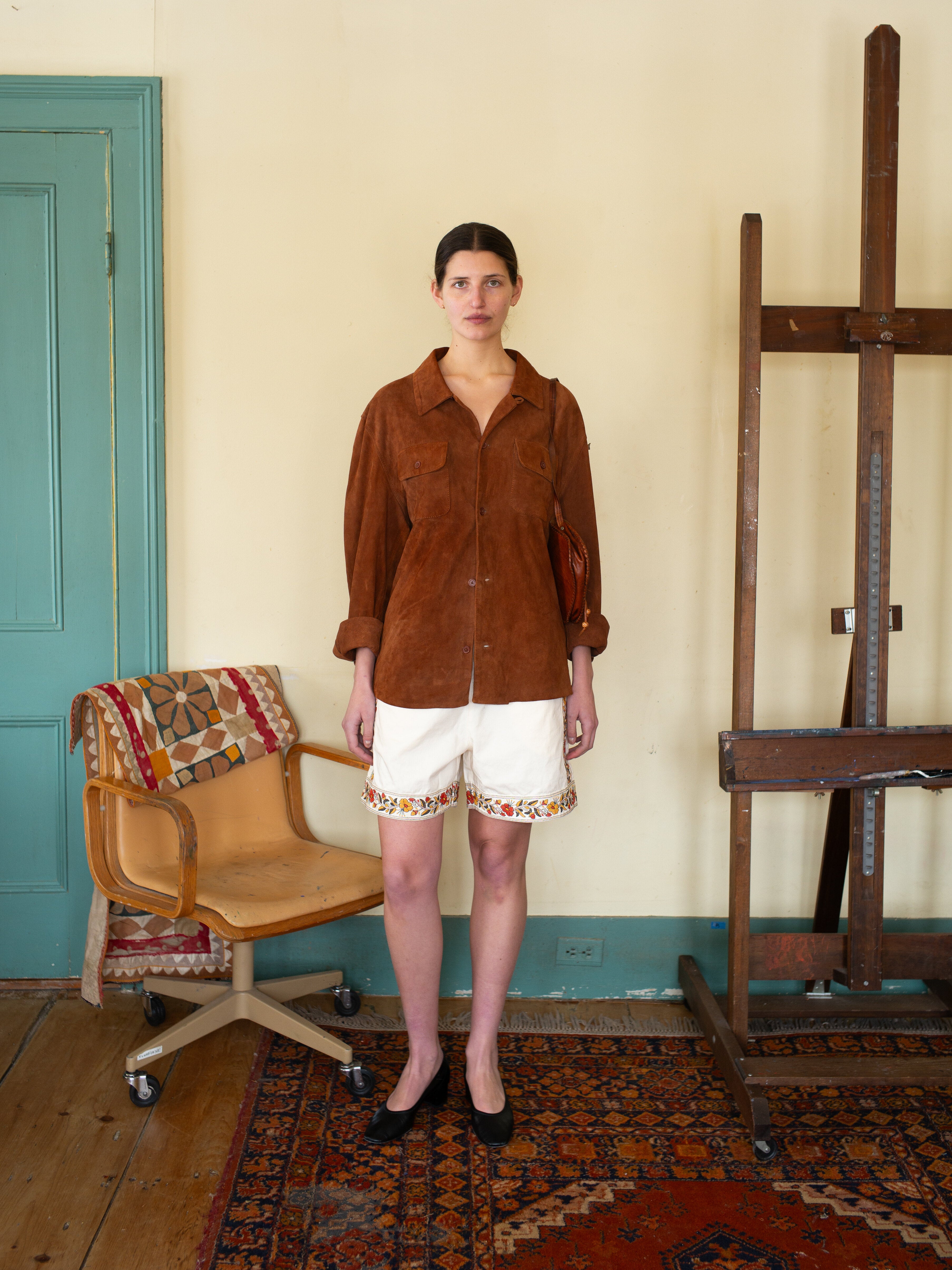 A person stands in a room with a wooden easel, wearing FOUND's rugged workwear-inspired Suede Camp Shirt and white shorts. To the left is an empty beige chair with a colorful cushion, enhancing the inviting vibe that complements the creative ambiance.