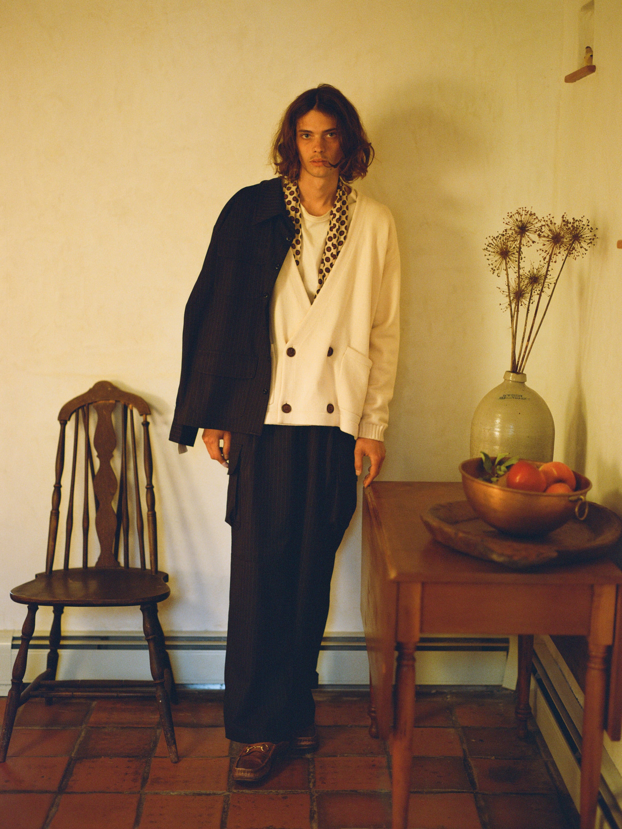 Someone with long hair wears FOUND's Double Breasted Knit Cardigan and loose pants, standing between a wooden chair and a table adorned with a vase and fruit bowl. The ensemble is perfectly complemented by the midweight cotton blend attire.