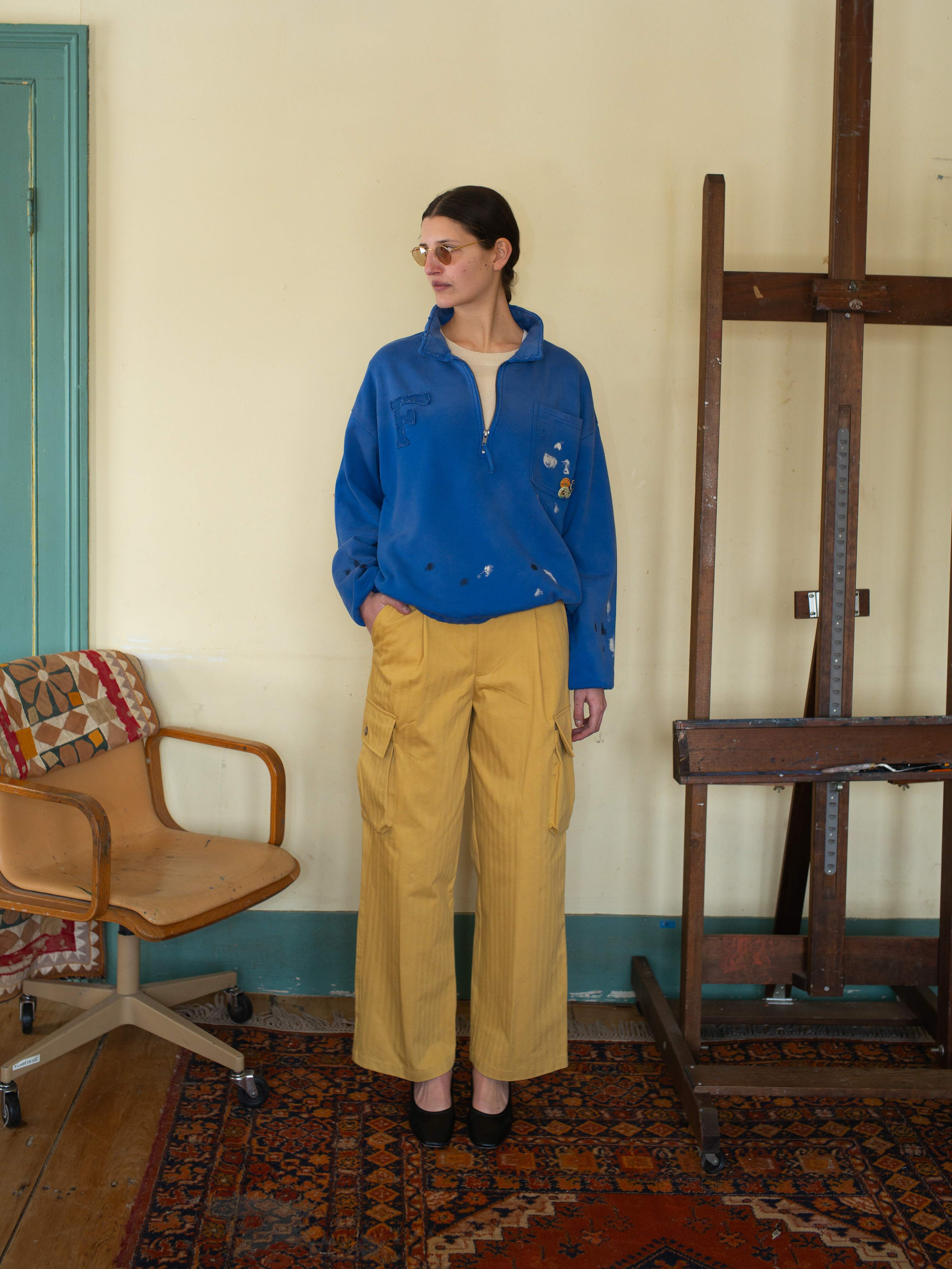 A person wearing a FOUND Painters Pullover Sweatshirt and yellow cargo pants stands beside an easel and an office chair on a patterned rug, blending style with workwear.