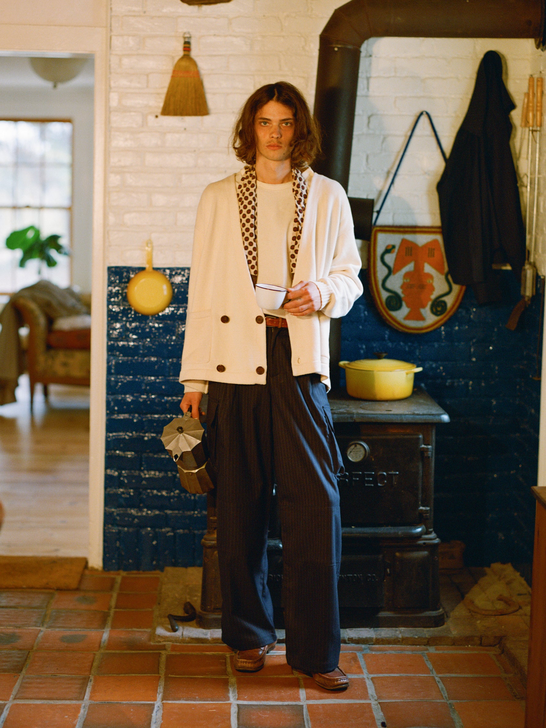 In a kitchen, a person with shoulder-length hair wears a white jacket while holding a cup and kettle. Their style shines with FOUND's Hava Pinstripe Pleated Cargo Trousers, made from wool-blend fabric and featuring practical cargo pockets. A wood stove and cookware complete the backdrop.