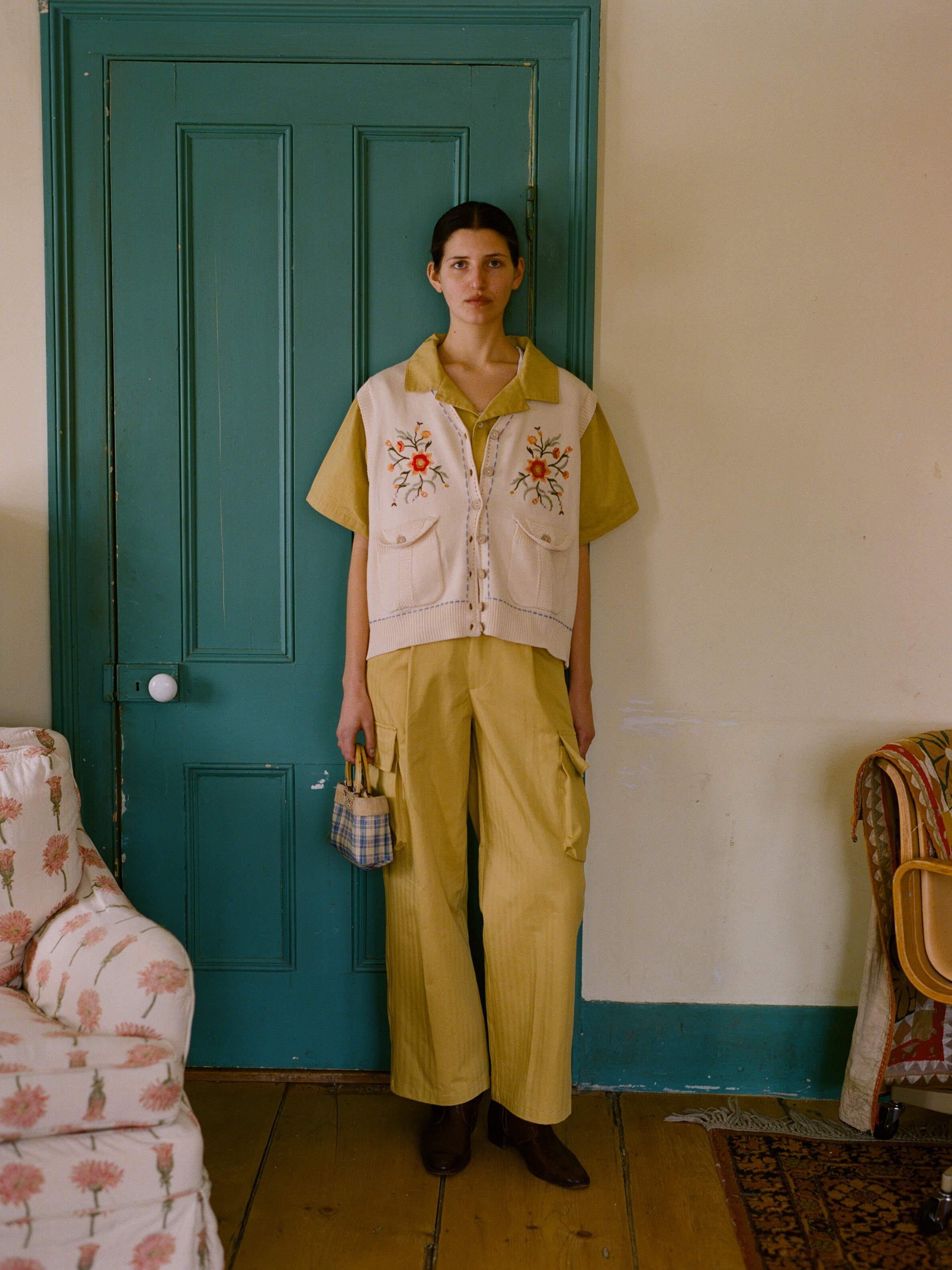 In front of the green door, wearing a relaxed fit yellow outfit with a floral vest and holding a plaid bag. This includes FOUND's Sunlight Twill Cargo Pants for versatility. A patterned couch is on the left, and a wooden chair is on the right.