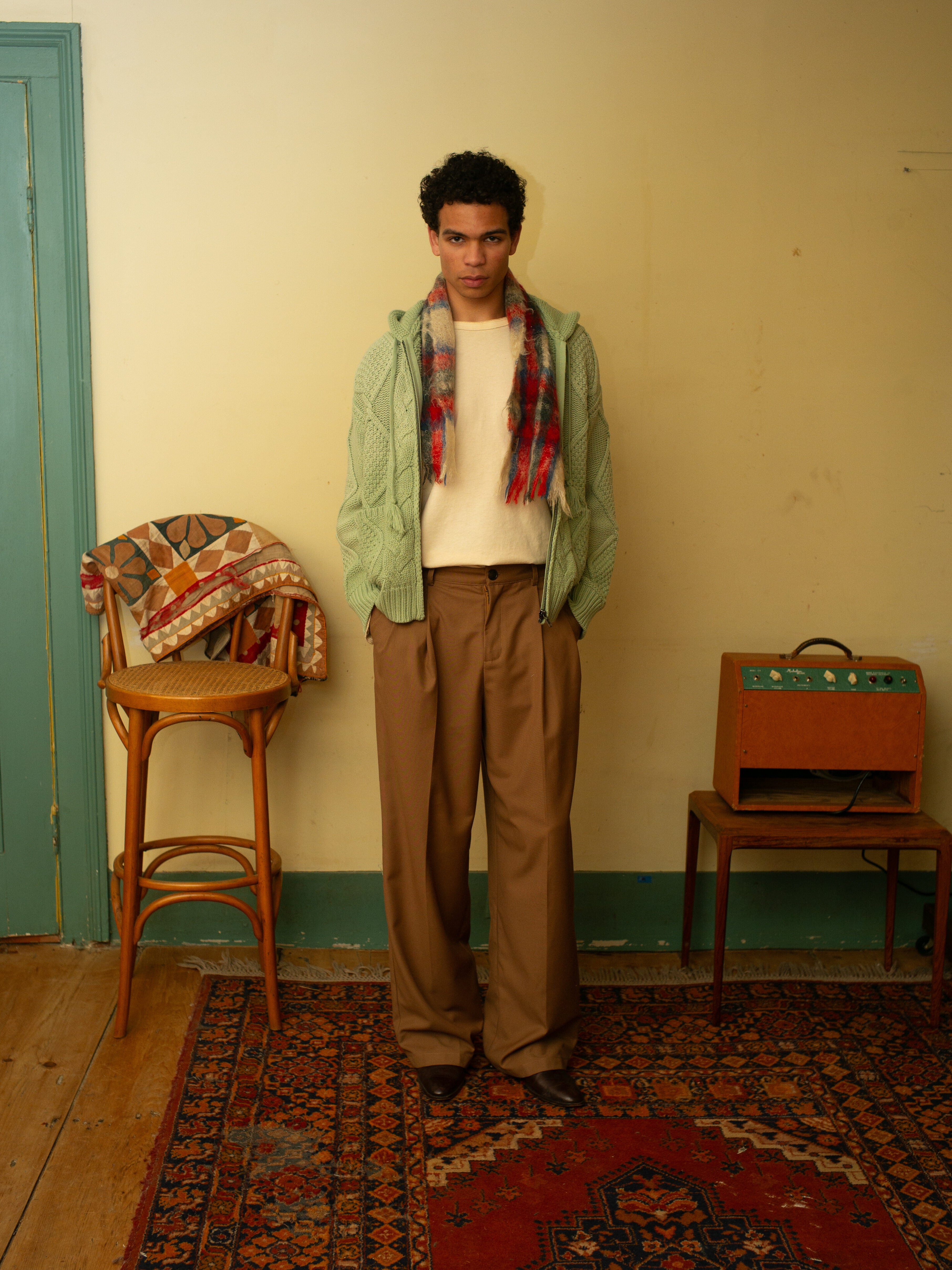 A person stands in a room wearing FOUND's Zip Up Cable Knit Hoodie, a white shirt, plaid scarf, and brown pants. Nearby, a wooden chair with patterned cloth sits beside a small electronic device on the floor.