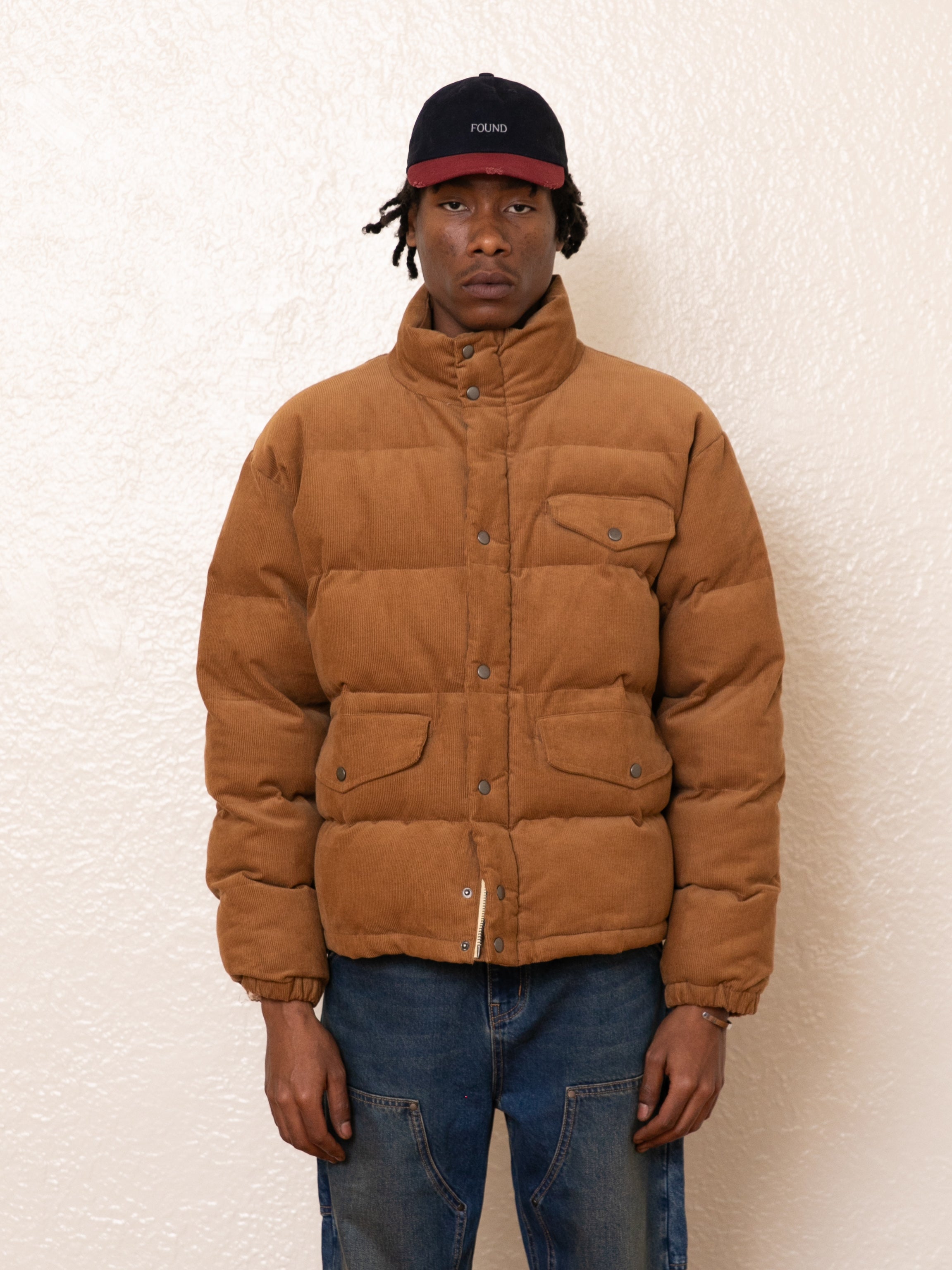 A person wearing a tan FOUND Corduroy Puffer Jacket, blue jeans, and a black cap stands against a textured light-colored wall.