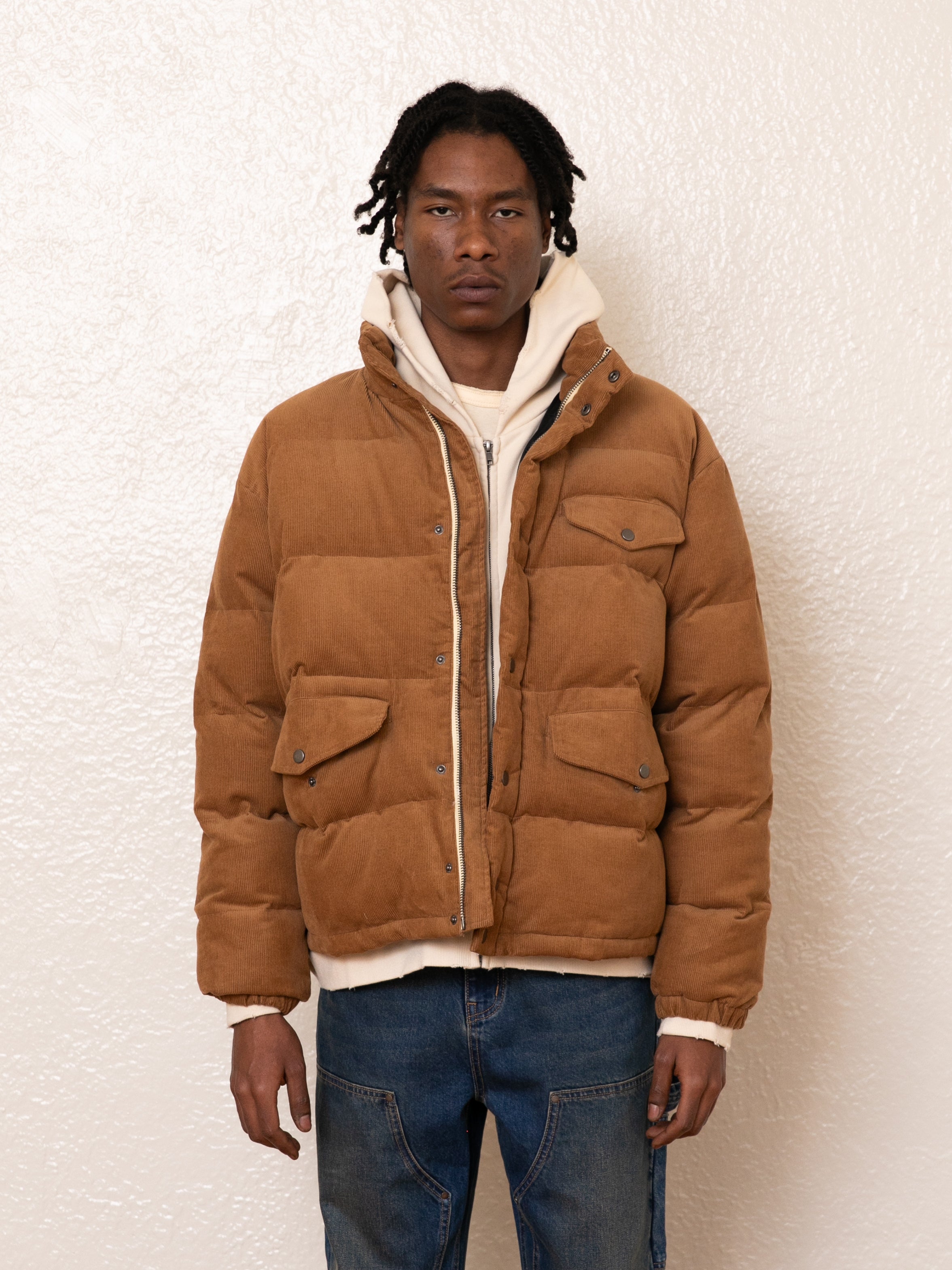 A person wears a warm tan Corduroy Puffer Jacket by FOUND over a white hoodie and blue jeans, standing against a plain white background.