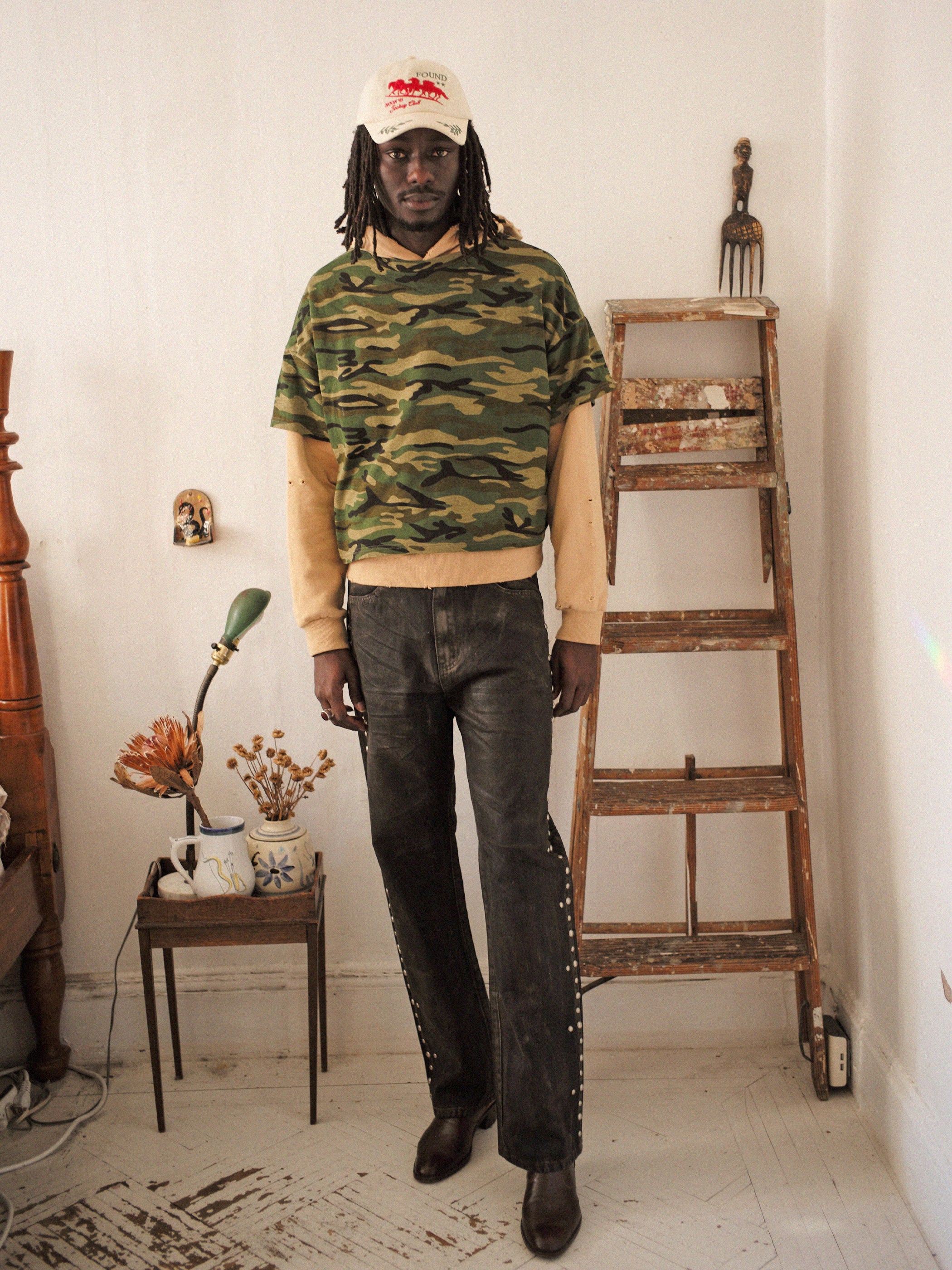 A person in a FOUND Double Layer Hoodie and jeans stands near a wooden ladder in a room with a small table holding dried flowers and a cup, wearing a baseball cap.