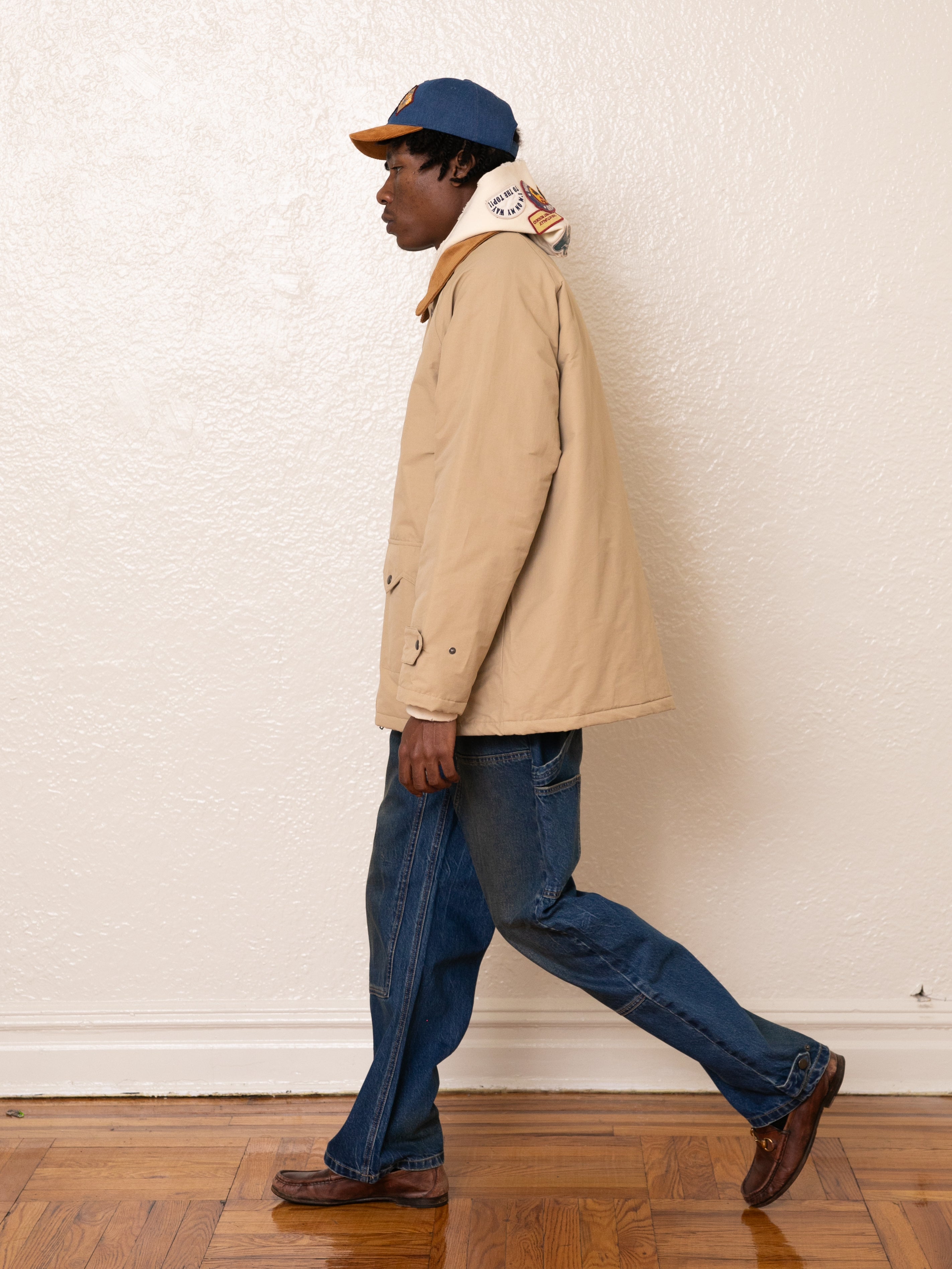 The person walks indoors wearing a FOUND Fisherman Jacket in beige with antique silver clasps, complemented by blue jeans, a blue cap, and brown shoes.