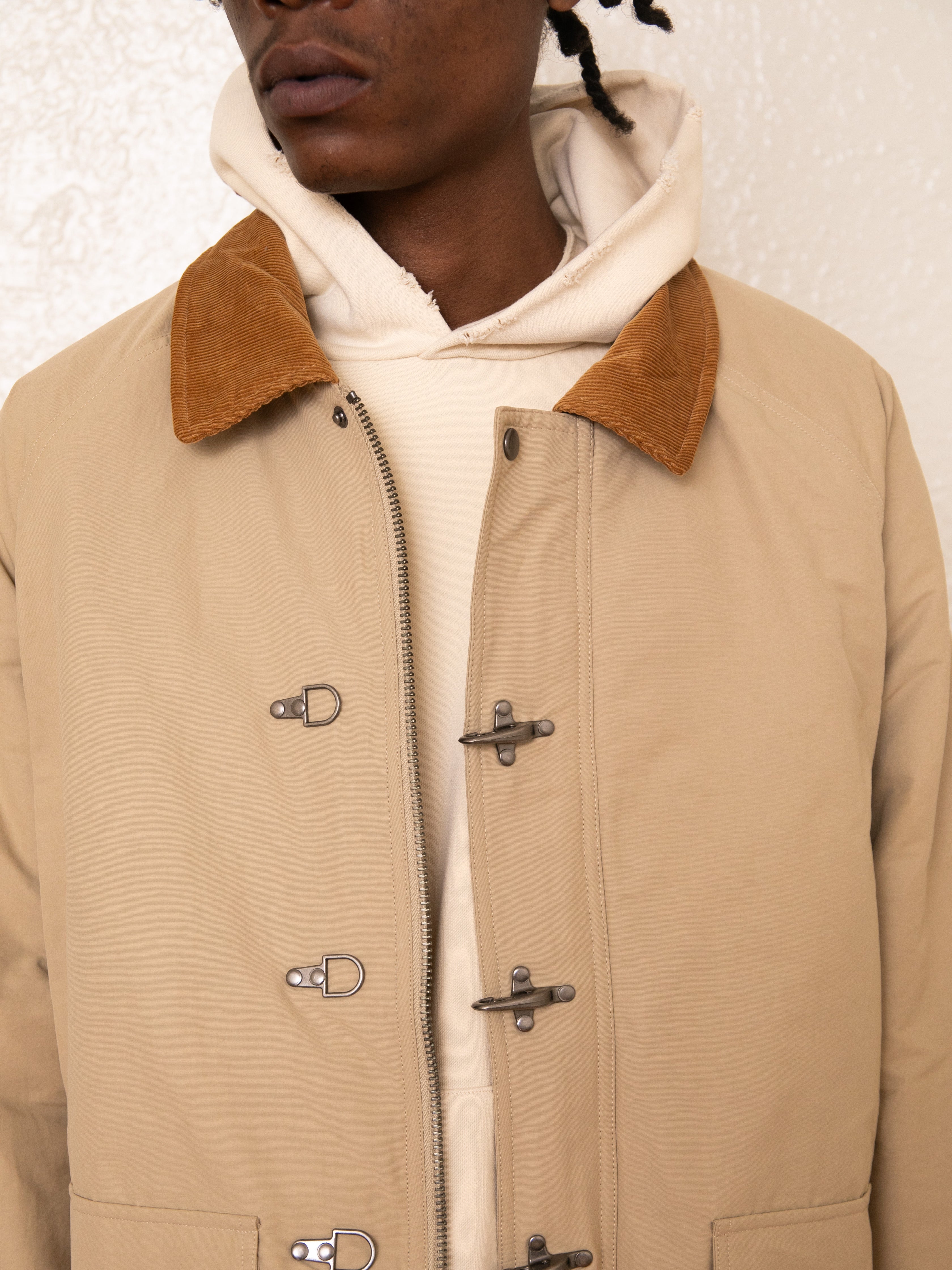 A person is wearing FOUND's Fisherman Jacket in tan, featuring a corduroy collar and toggle buttons, over a cream hoodie.