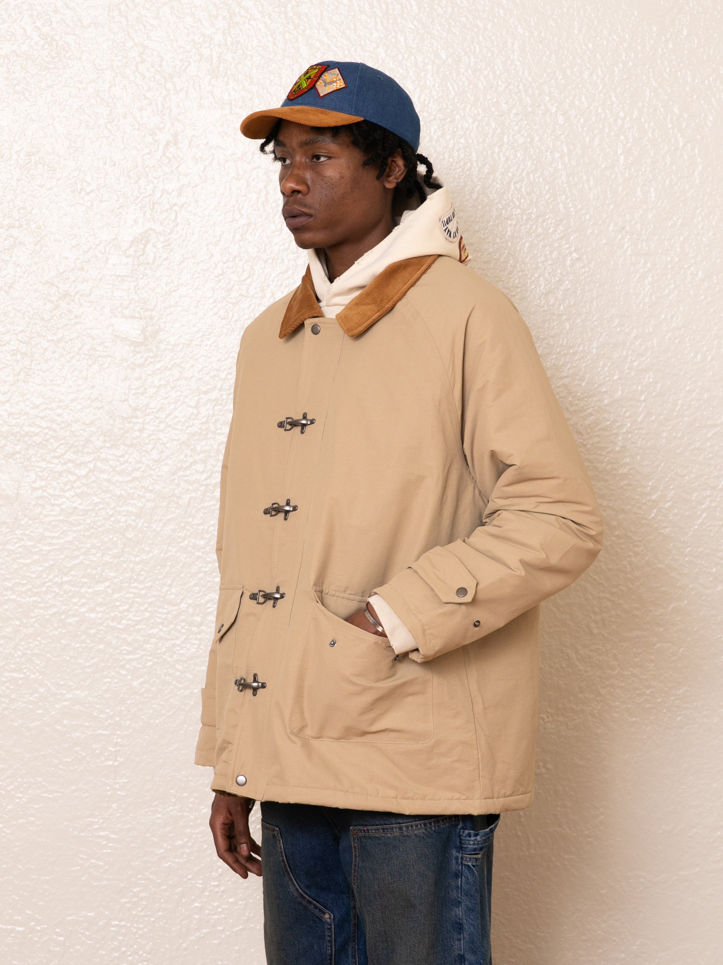 A person wearing a FOUND Fisherman Jacket with antique silver clasps, a white hoodie, blue jeans, and a blue cap stands against a textured white wall.