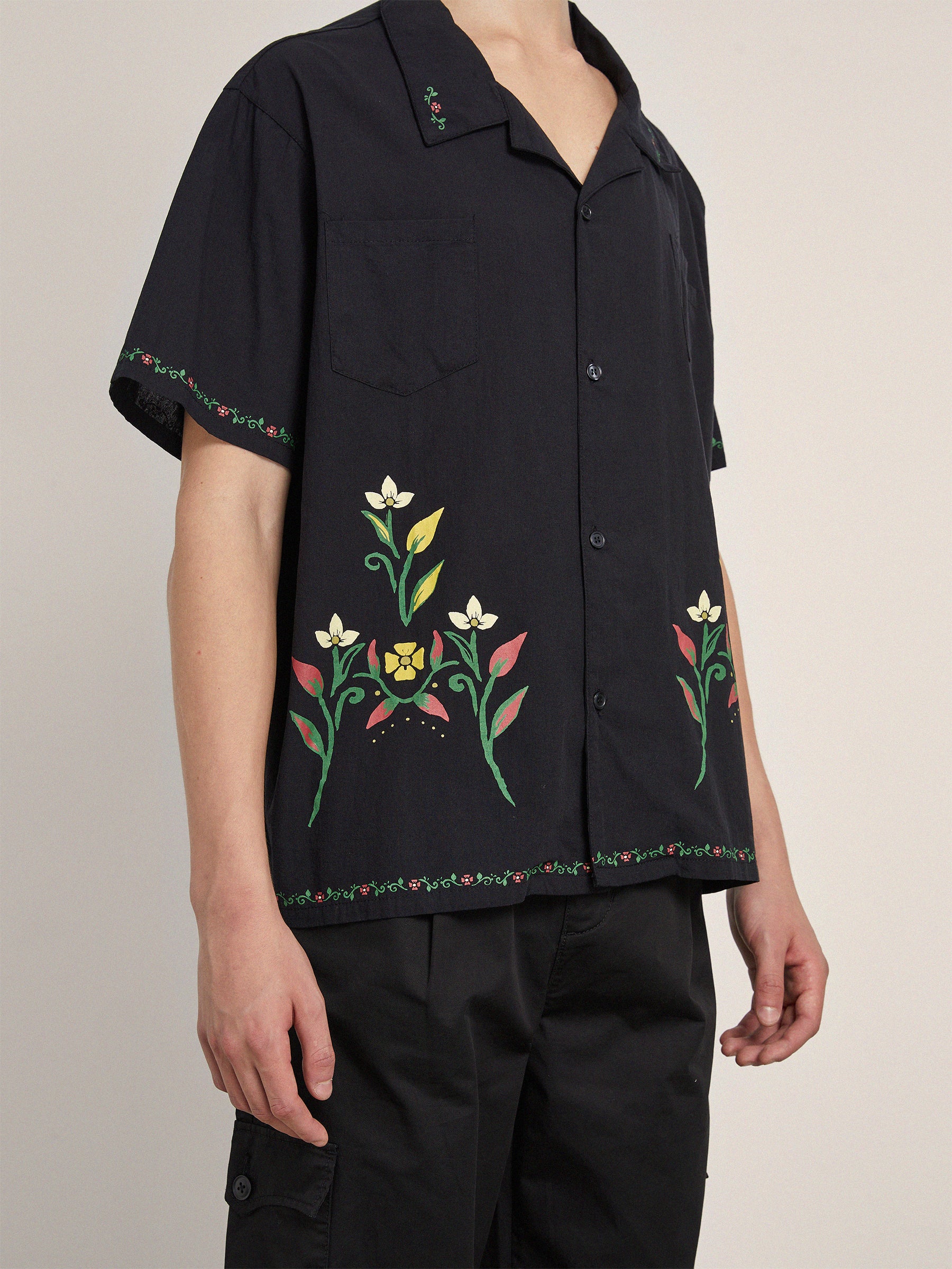 A person is wearing a black Profound Floral Camp Shirt, short-sleeve with floral embroidery on the front, sleeves, and hem. Made from 100% cotton fabric for a relaxed fit, they pair it with black pants. The background is neutral.