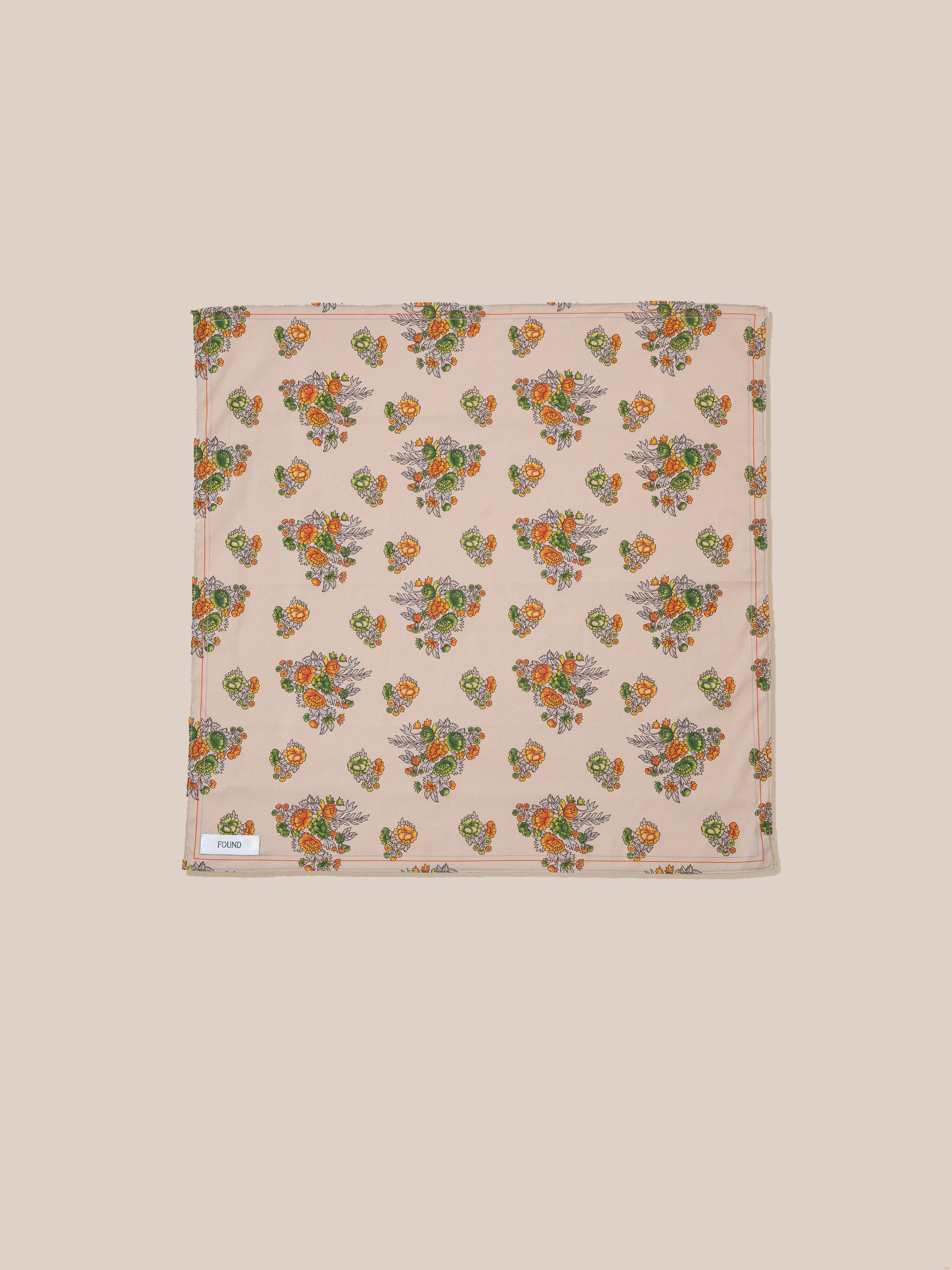 The Floral Print Bandana by Found features a square fabric with a beige background adorned with a Phulkari-inspired floral pattern in green and orange. A small tag with text is sewn onto one of the edges.