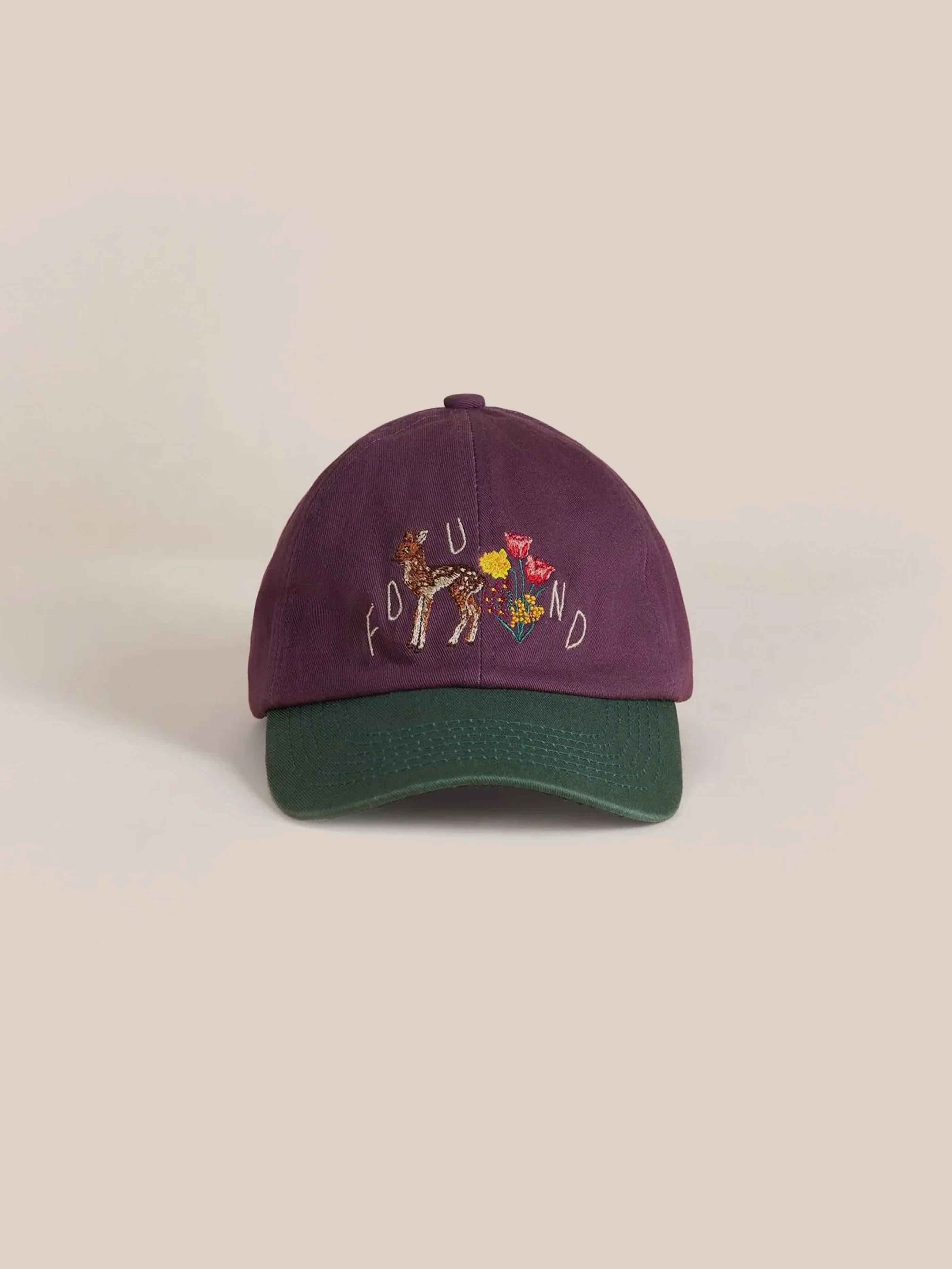 The Flower Deer Cap by Found is a purple and green baseball cap adorned with embroidered letters and images on the front, showcasing intricate floral chainstitching inspired by American folk art.