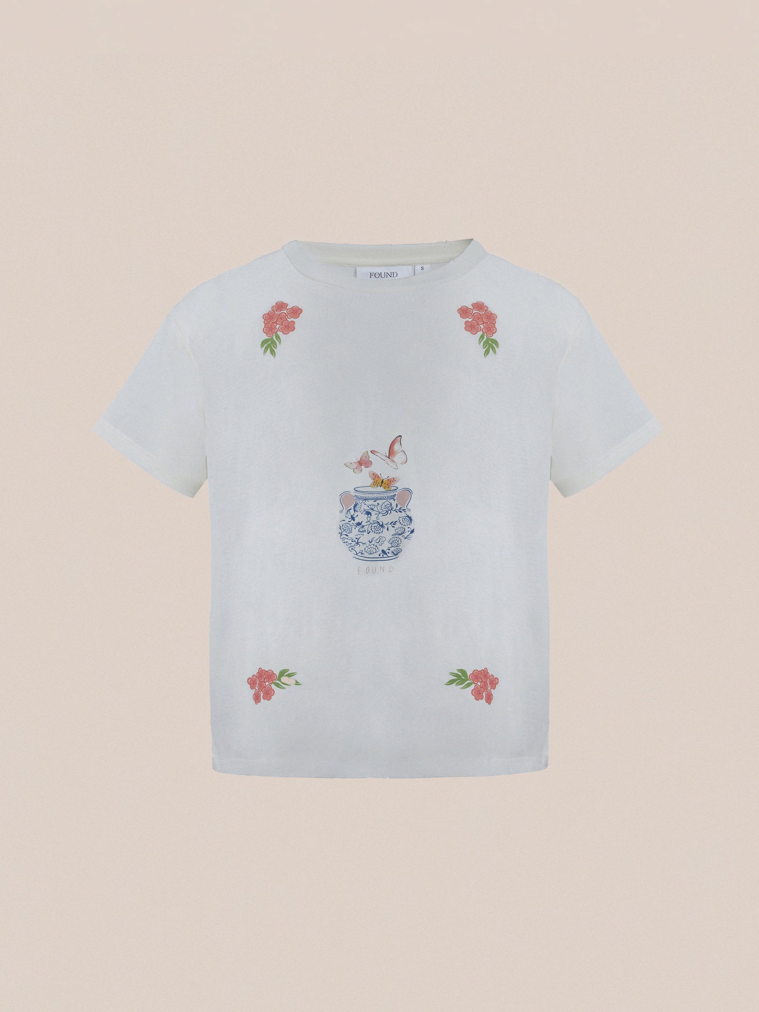 The Flower Pot Tee by Found is a white T-shirt adorned with floral accents and a whimsical design of two birds perched on a decorative vase, exuding springtime charm.