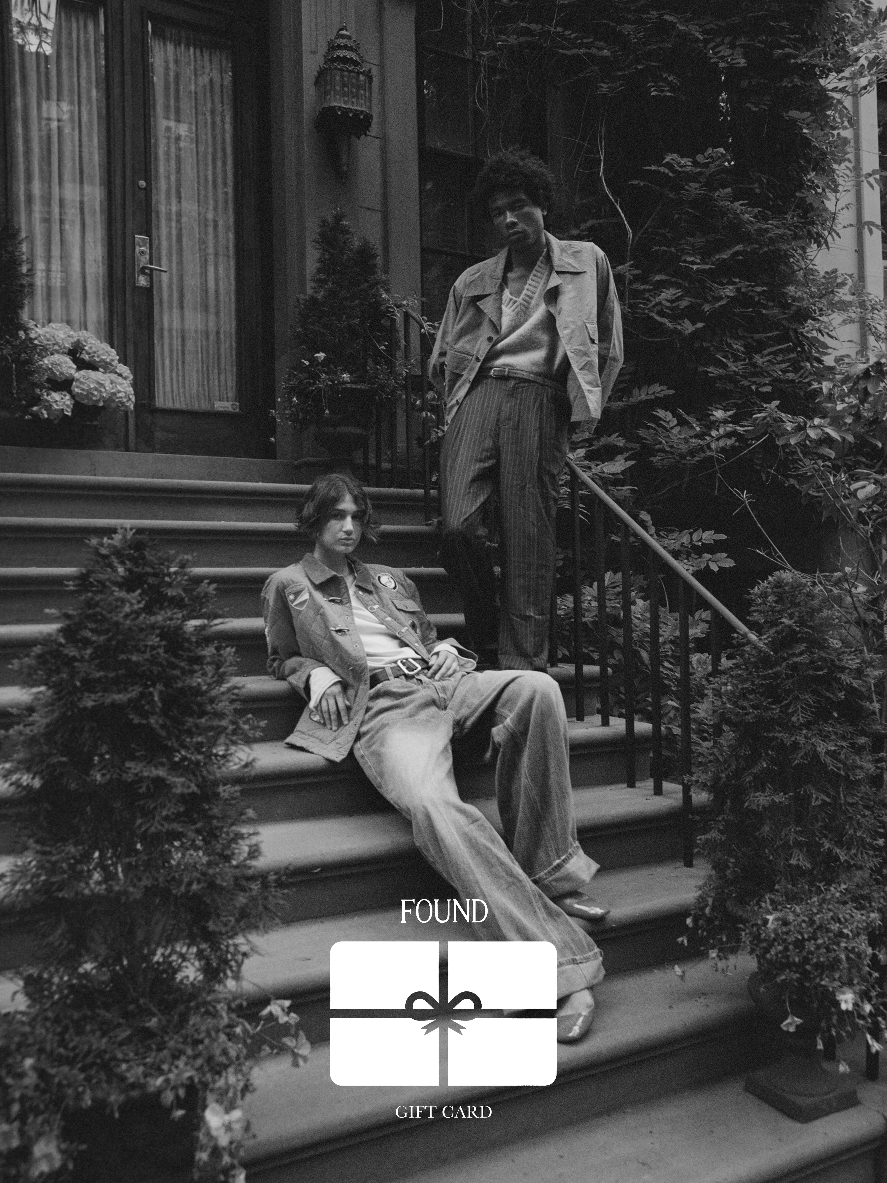 Two individuals in chic attire stand on a staircase outside a building covered in vibrant plants. In the center, an elegant graphic of the Found Gift Card by FOUND is prominently displayed. The black and white image exudes an air of enduring sophistication.