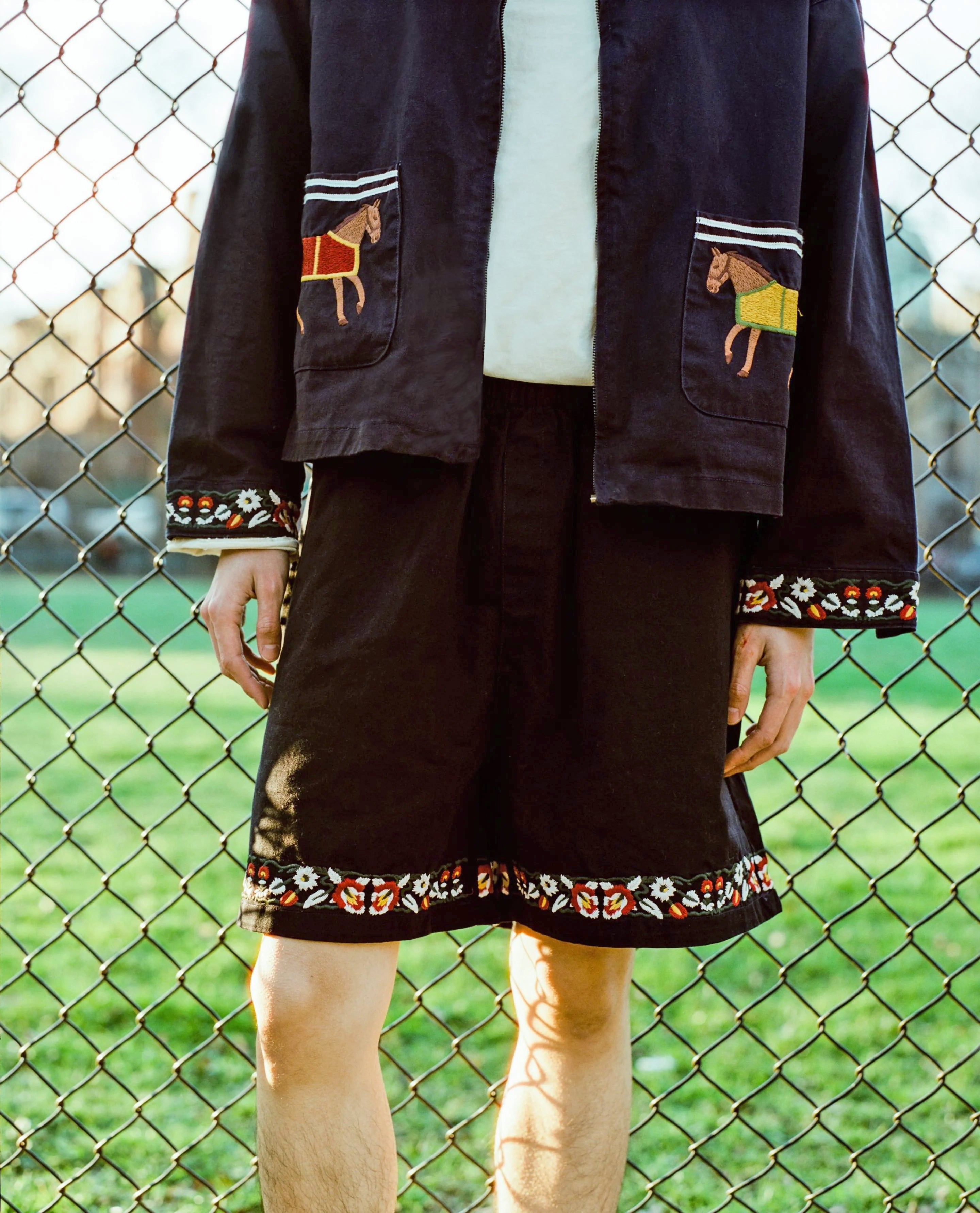 A person wearing a detailed black jacket with floral embroidery and Found Horse Equine Twill Shorts stands behind a chain-link fence, with sunlight casting shadows on the ground.