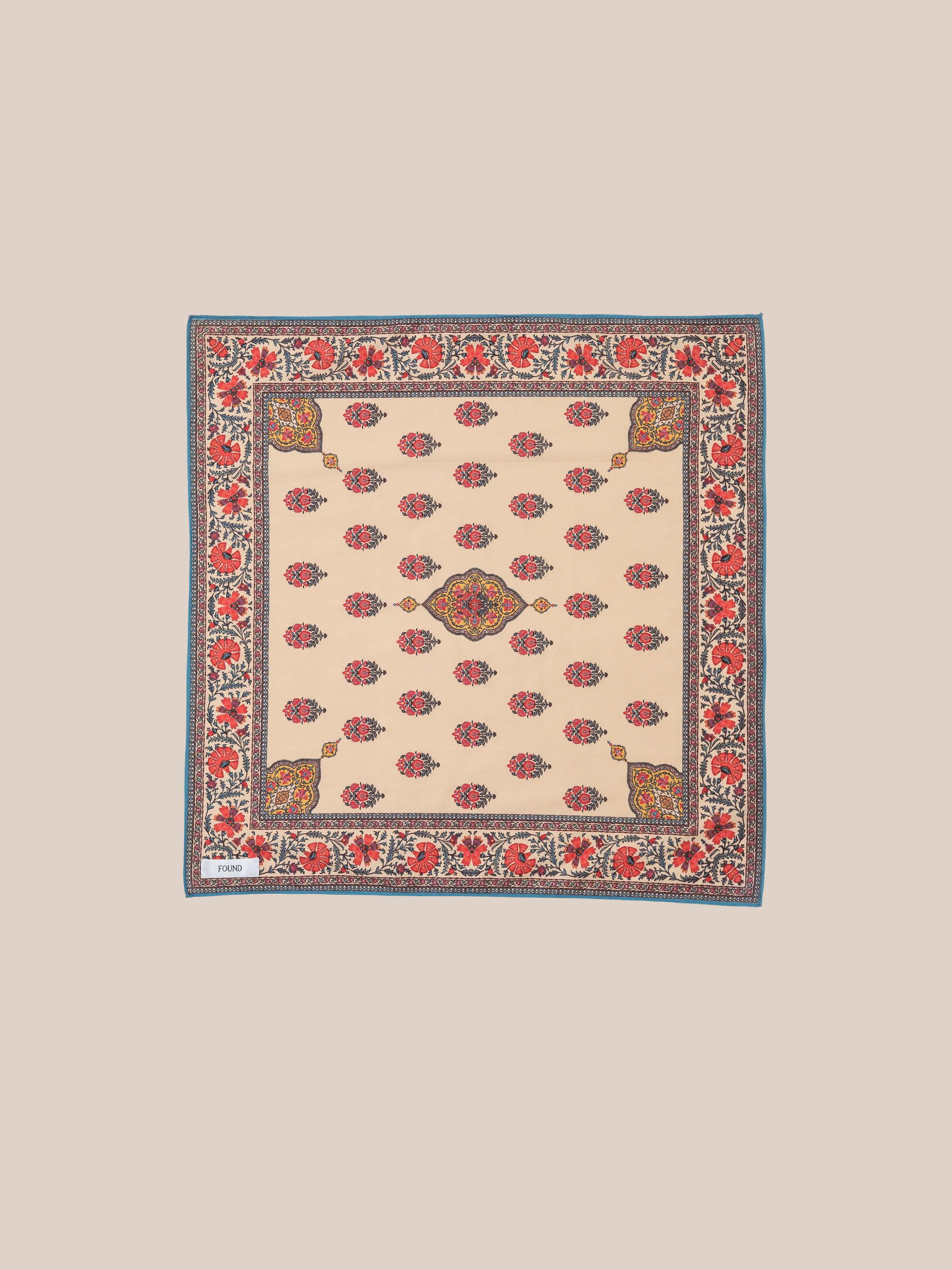 The Poinciana Bandana by Found is a square scarf made from a cotton blend, displaying an intricate floral pattern in red and blue on a beige background. Its border highlights an elegant floral design with a blue trim, inspired by traditional Indo-Aryan prints.