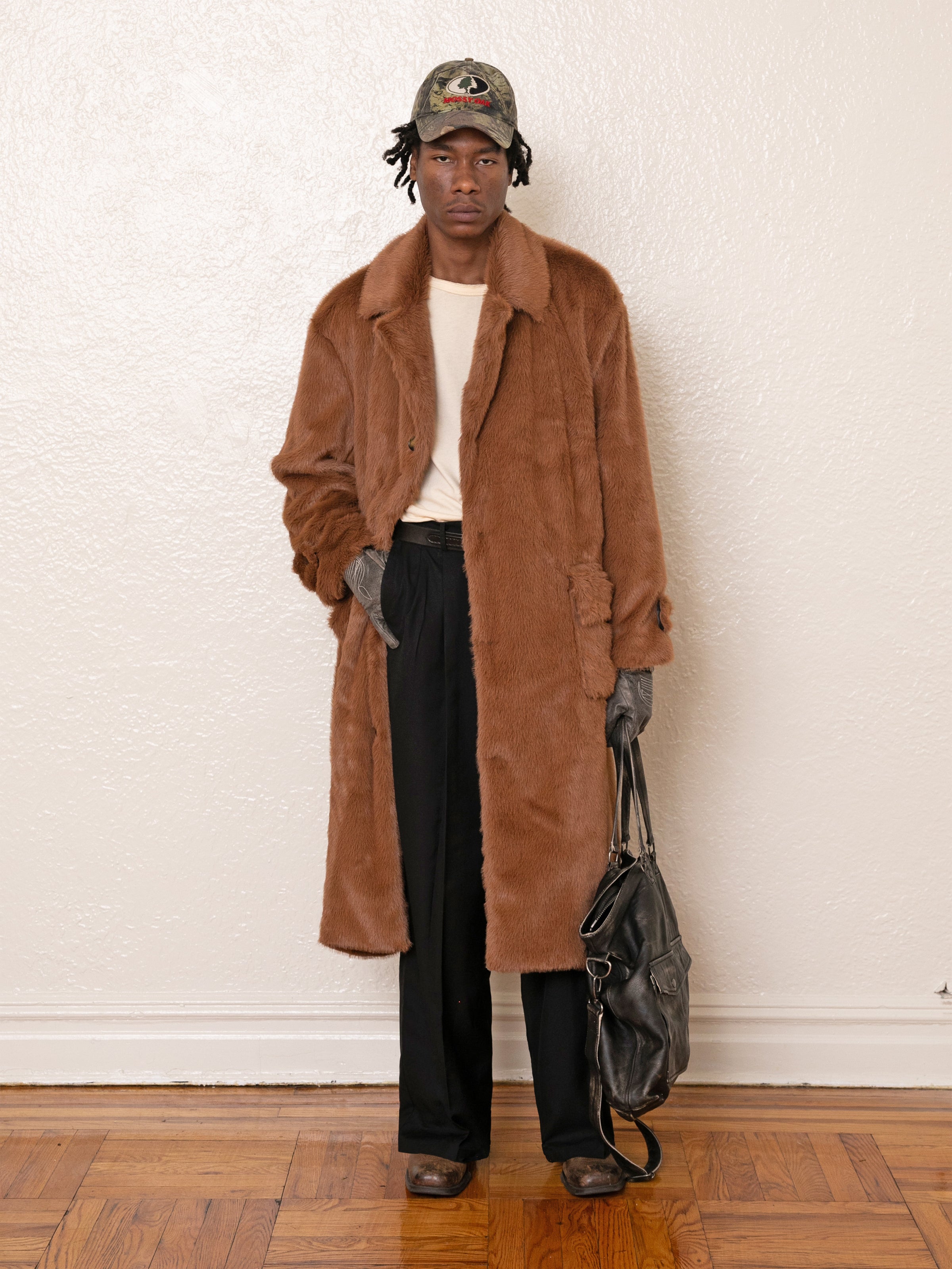 A person stands against a white wall wearing a FOUND Long Faux Fur Coat, black pants, gloves, and a patterned cap, holding a black bag in one hand.