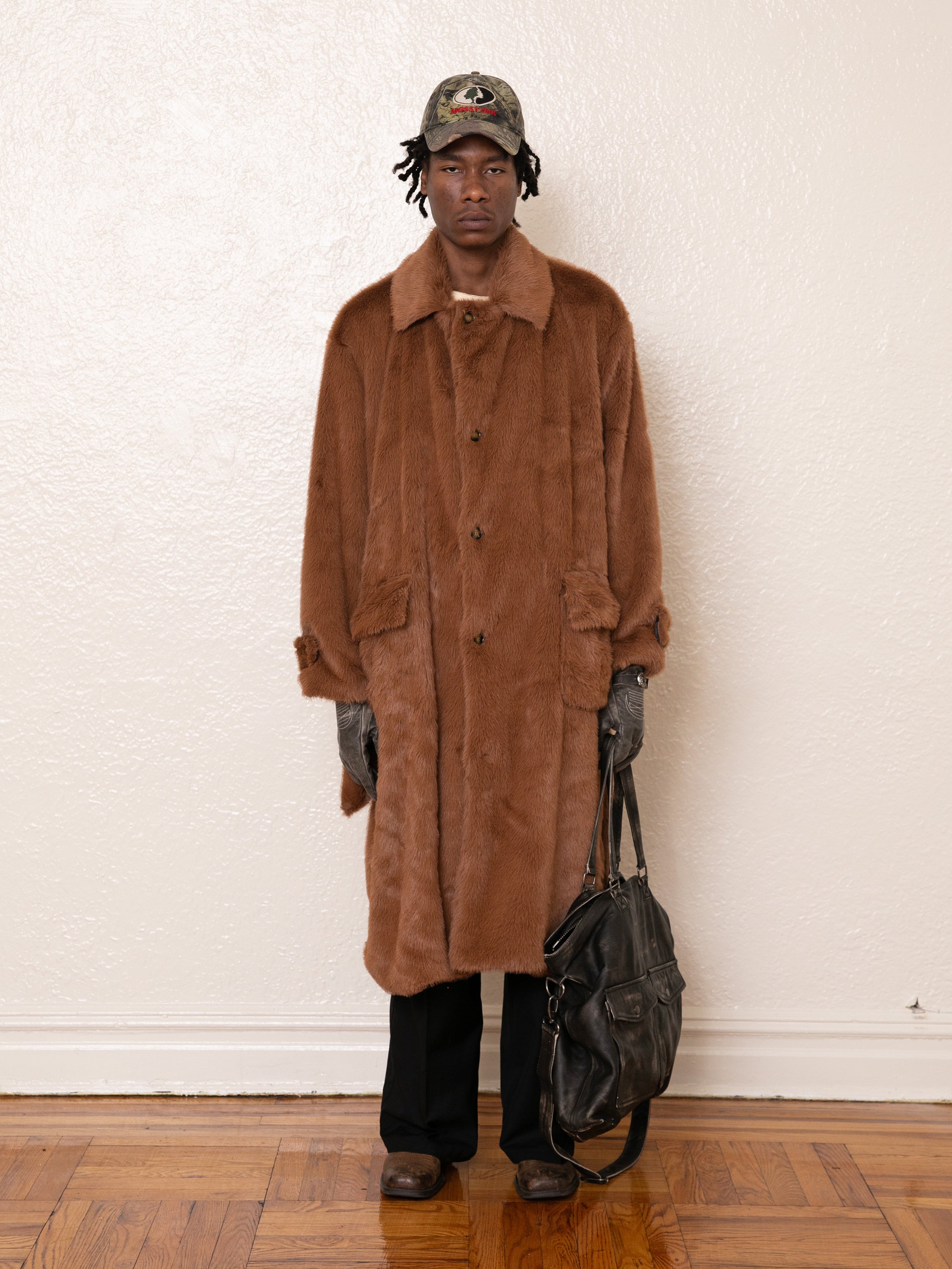 A person wearing a FOUND Long Faux Fur Coat and a hat stands holding a large black bag against a plain wall on a wooden floor.