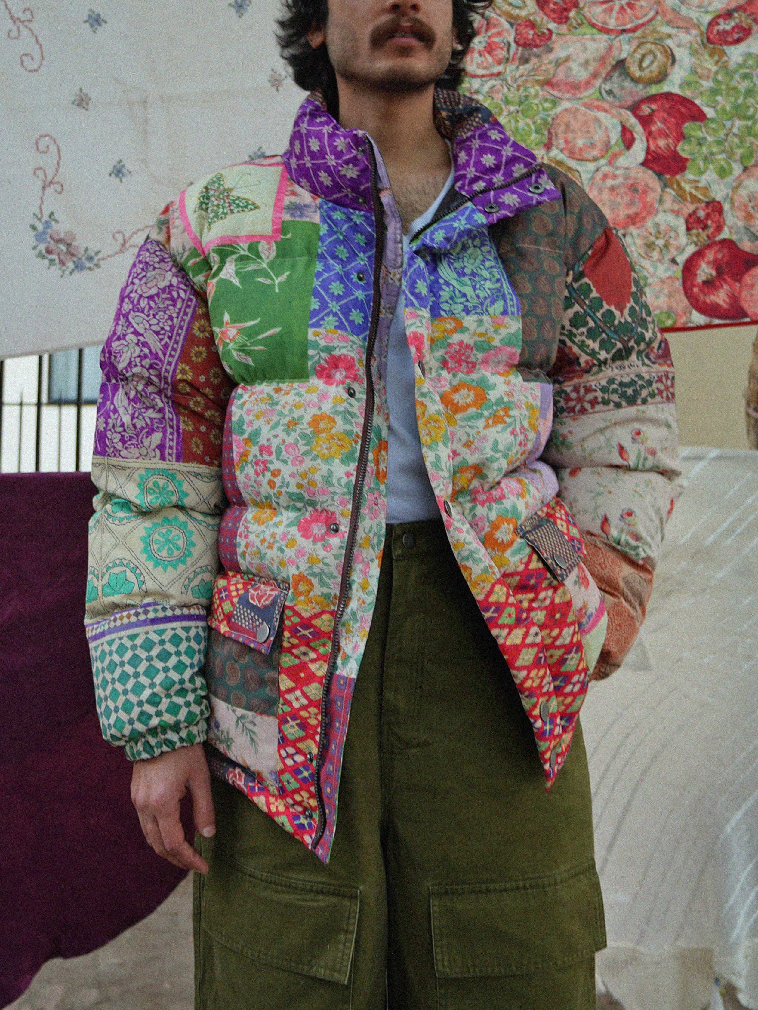 A person dressed in a vibrant Gardenia Tapestry Puffer Jacket by Found, paired with olive green pants, stands before a floral tapestry background, highlighting the elegance of South Asian prints.