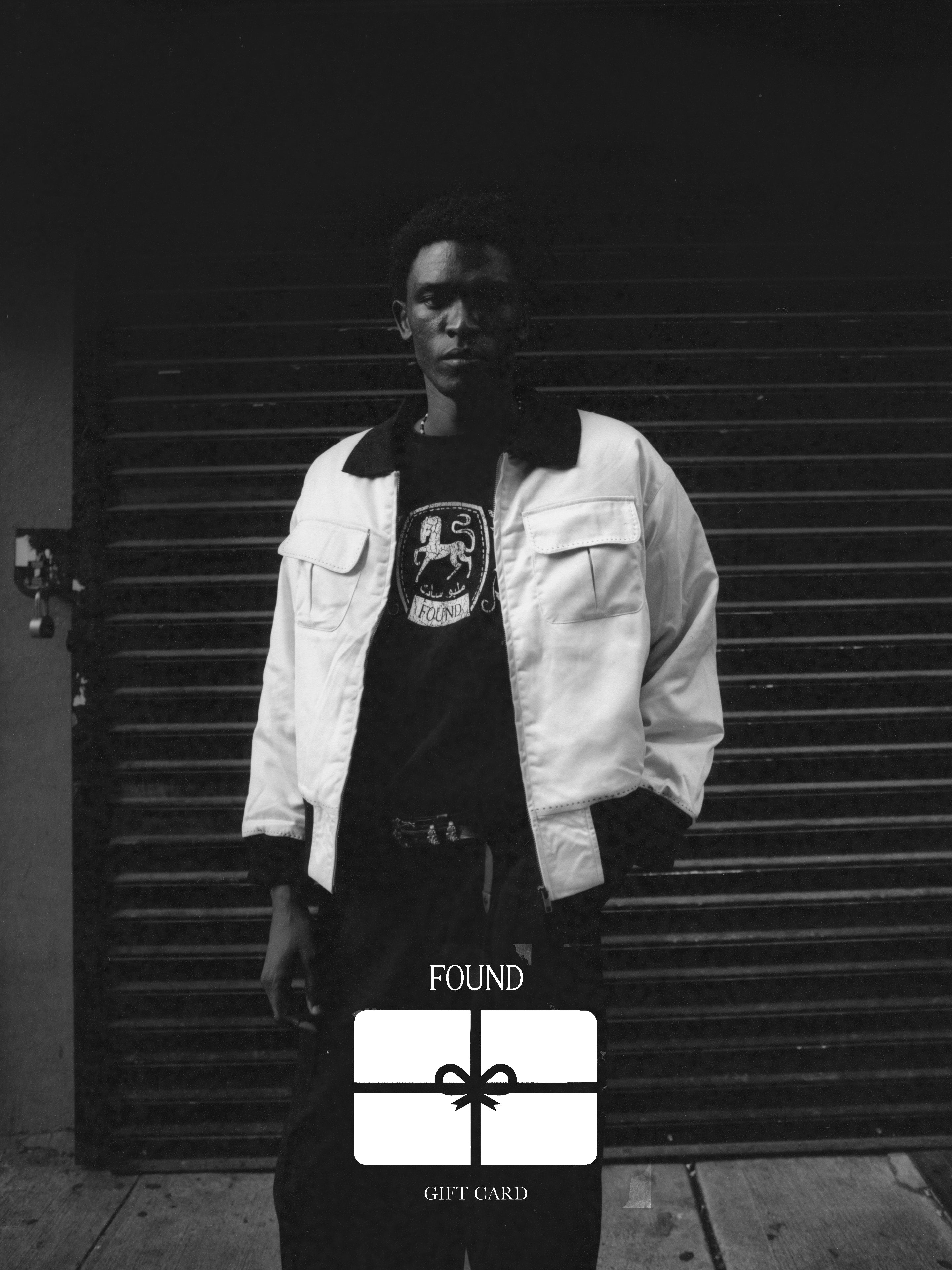 A person stands in front of a corrugated metal door, adorned in a white jacket and dark pants, emanating an air of sophistication. An elegantly designed "Found Gift Card" graphic is placed at the bottom of the image, showcasing the artistry associated with FOUND.