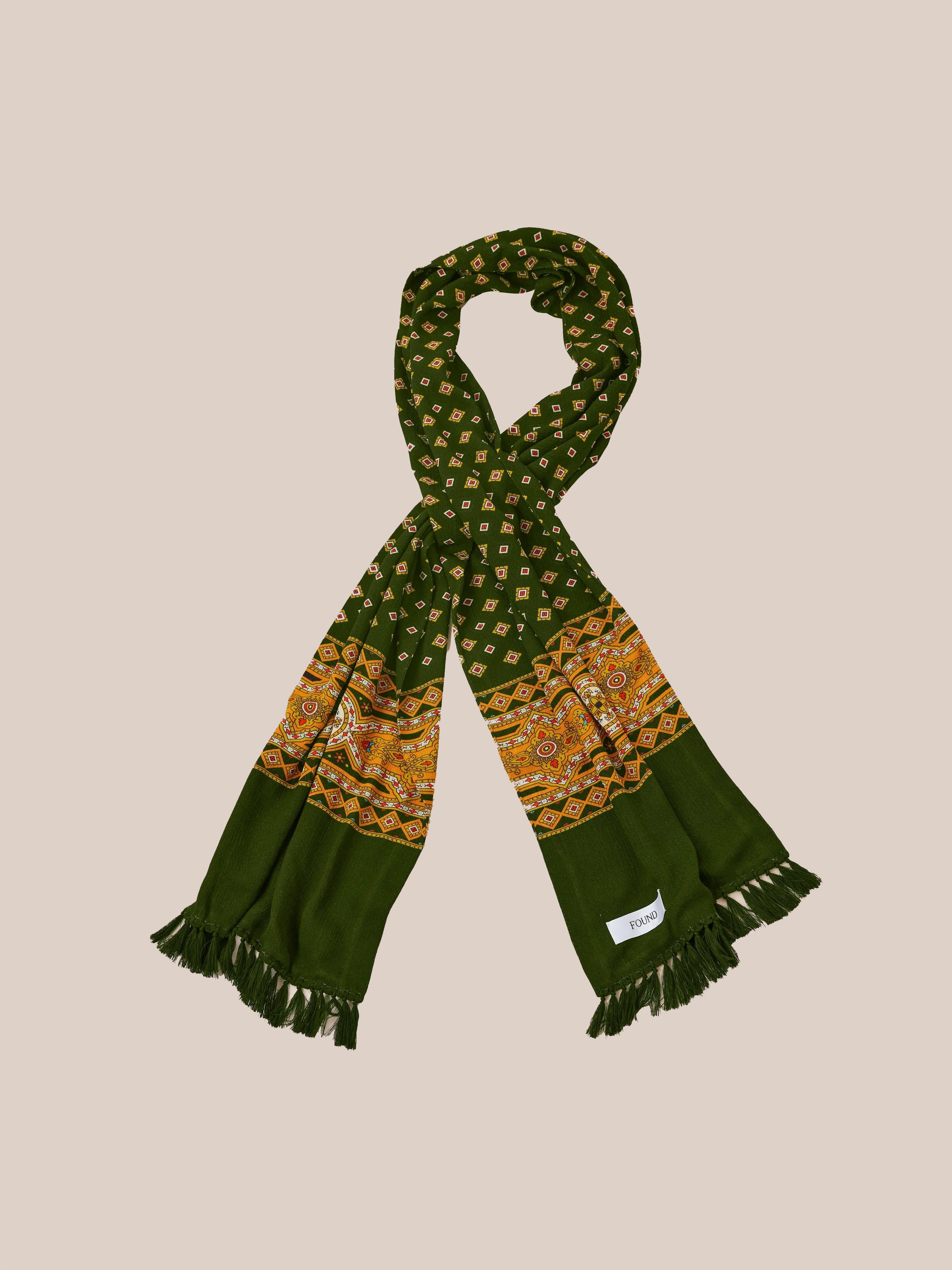 The Greener Pastures Scarf by Found boasts a vibrant geometric pattern adorned with ornate orange and gold designs at the ends, enhanced by hand-tied tassels. This exquisite accessory highlights Indo-Aryan prints, bringing cultural elegance to your wardrobe.