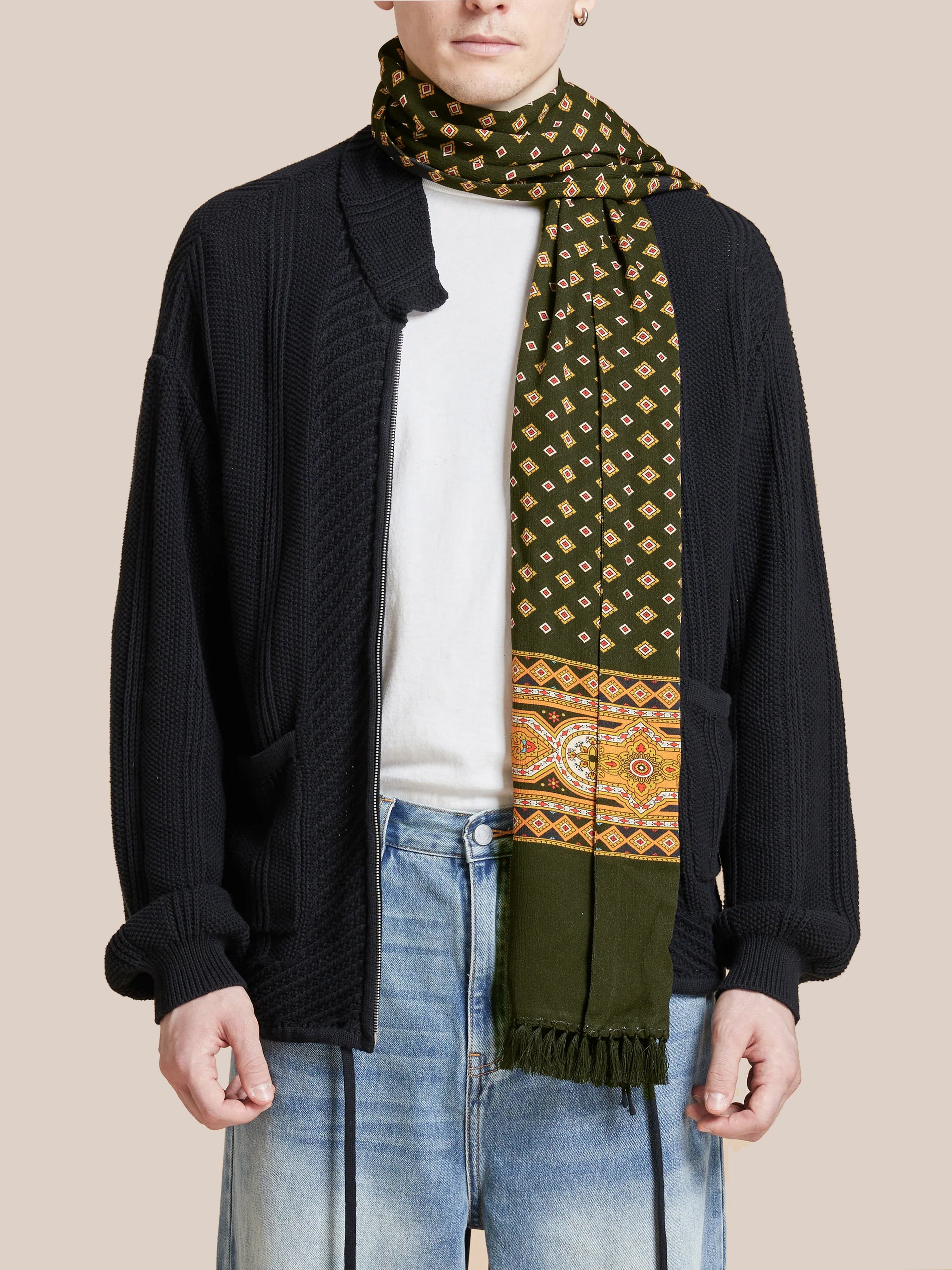 A person dressed in a black cardigan, white shirt, and denim jeans accessorized with a Greener Pastures Scarf by Found, featuring intricate Indo-Aryan designs, stands against a plain background.