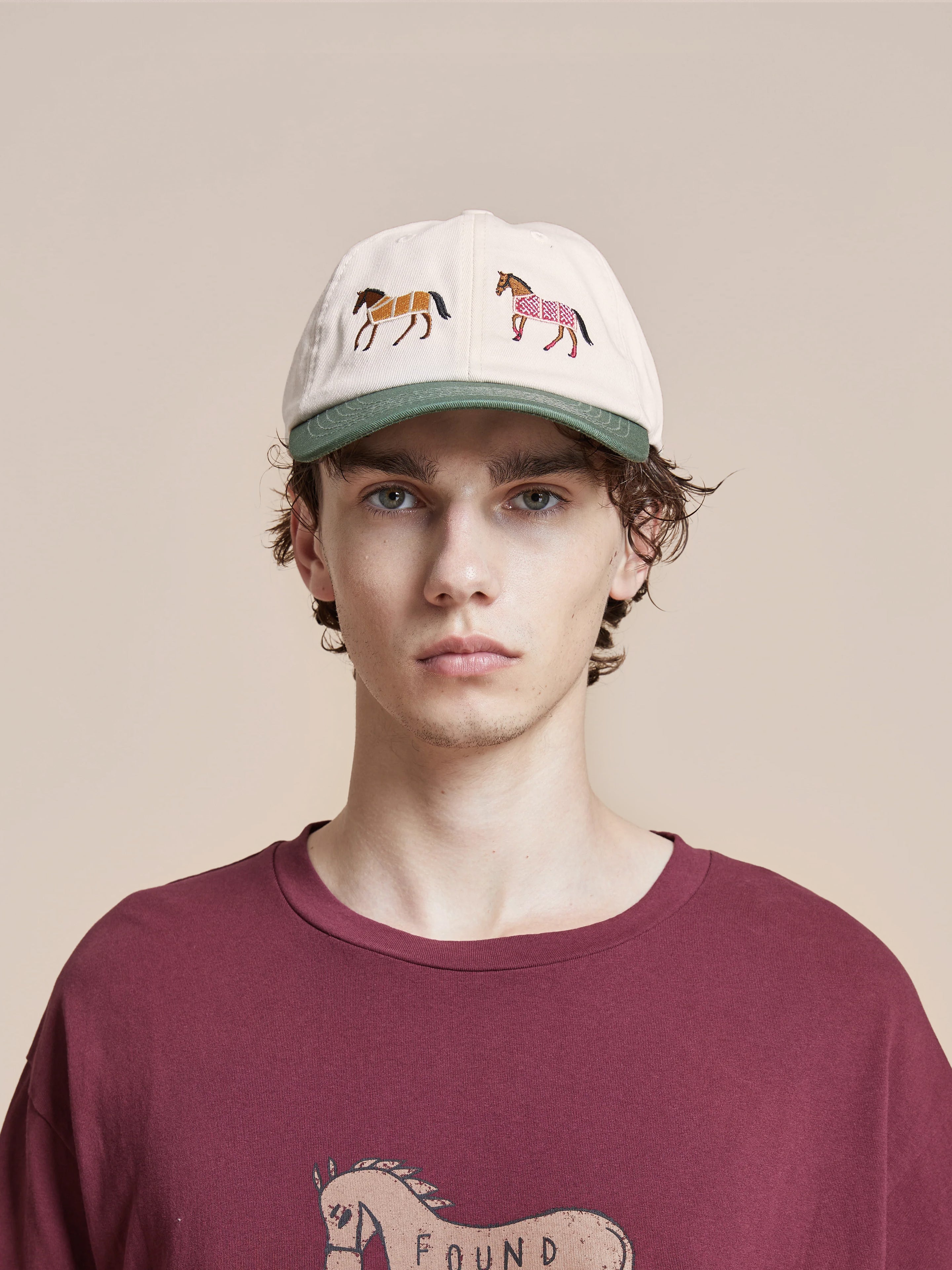 Wearing the "Horse Equine Cap" from Found, which features beige and green tones with intricate horse designs, and a maroon shirt adorned with a horse graphic, the individual stands against a beige background, epitomizing the spirit of racing colors.