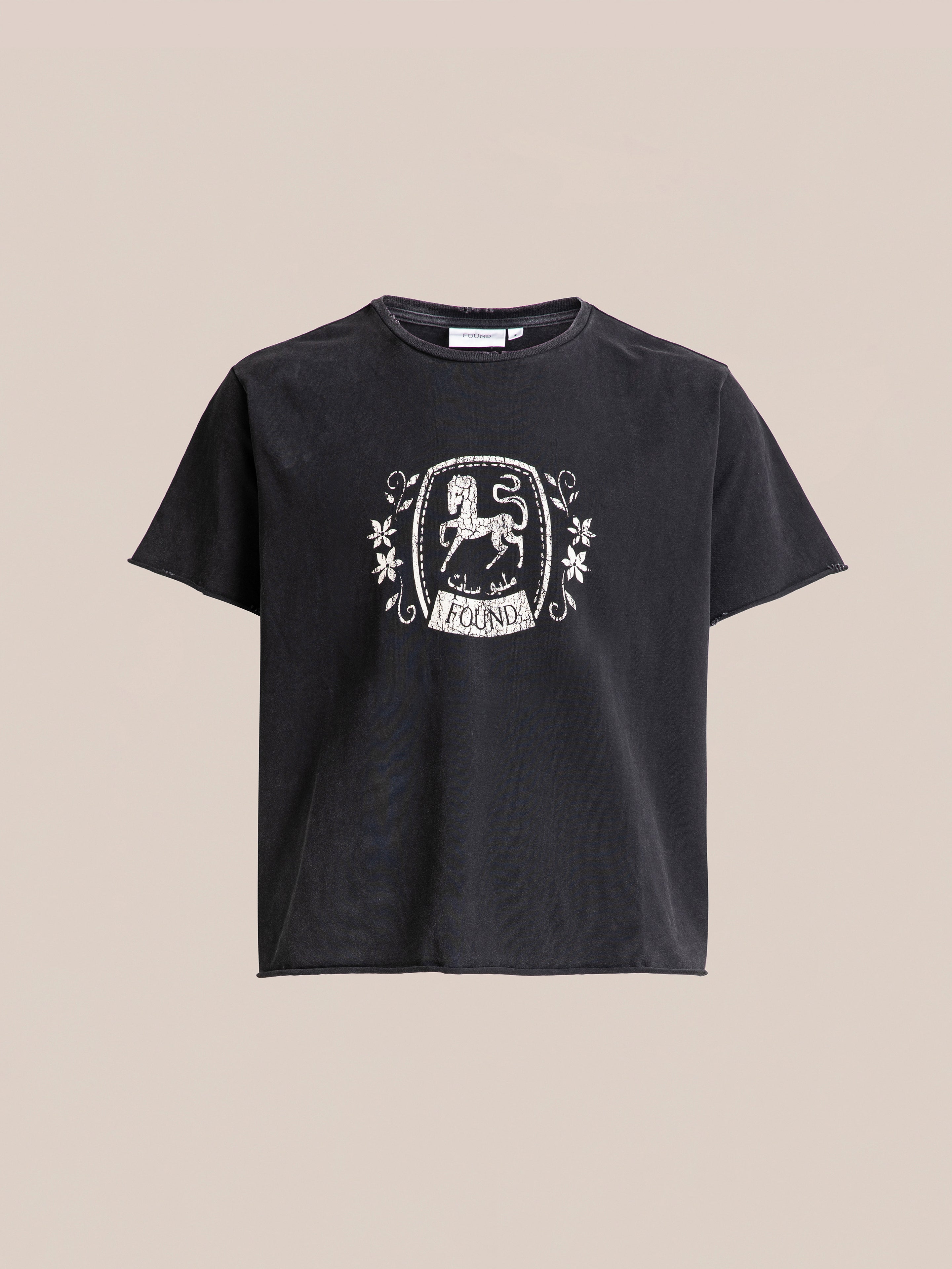 FOUND's Horse Logo Print Tee is a black T-shirt showcasing an intricate white print on the front, complete with decorative flourishes and a central crest design. Crafted from 100% cotton, it boasts an enzyme wash for added softness. The background is solid light beige.