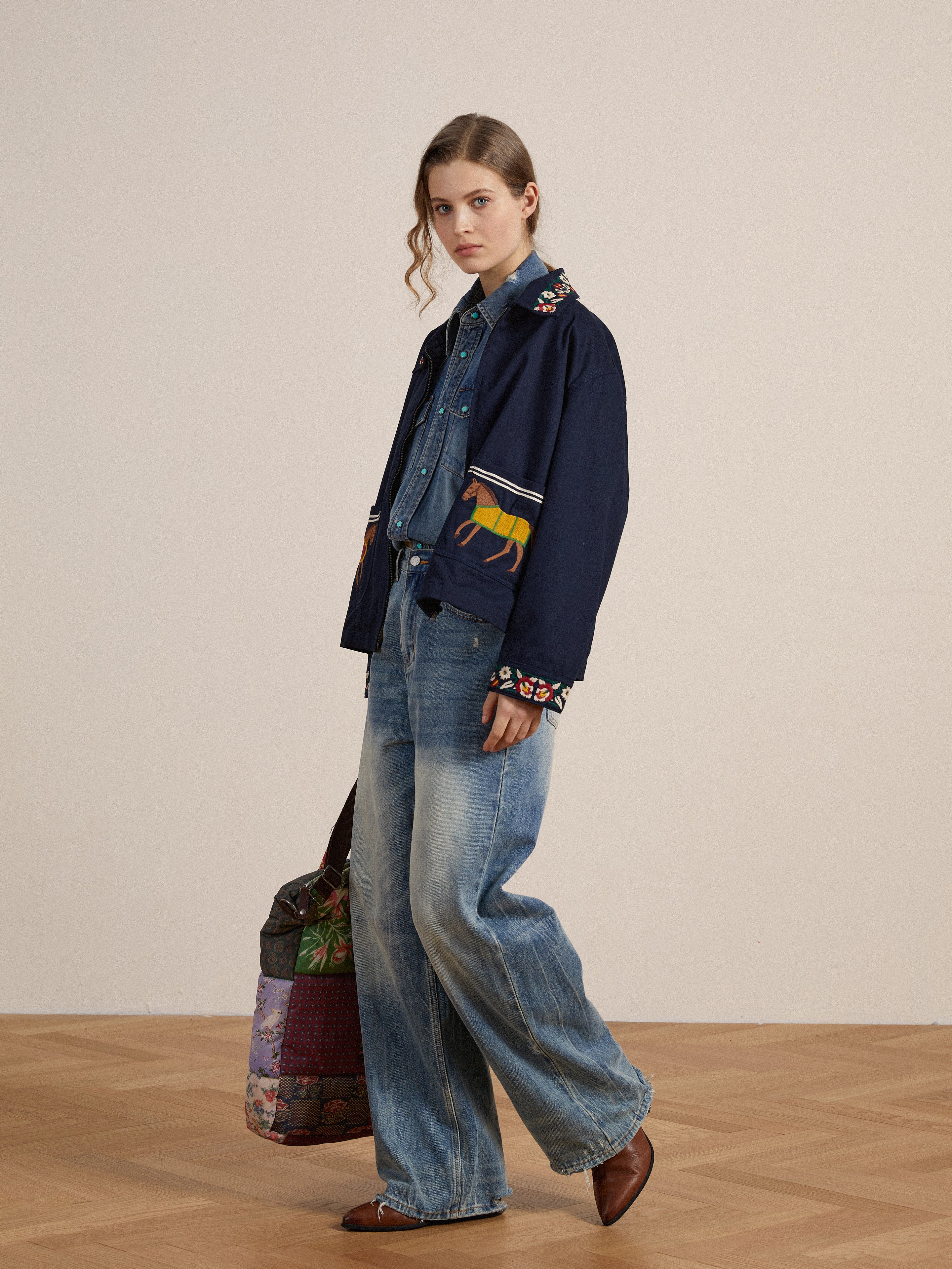 A woman in a Found navy blazer with detailed embroidery and denim jeans stands holding a patterned bag, against a neutral background.