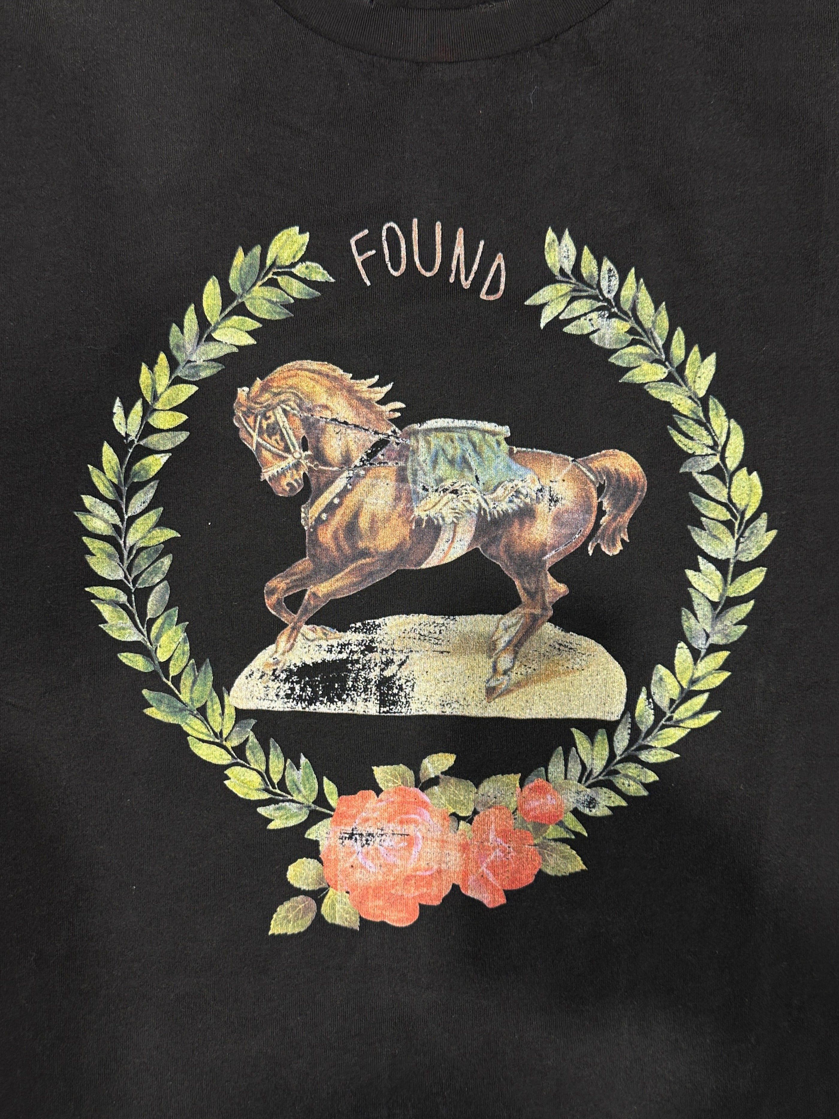 The Rodeo Tee by FOUND features an illustration of a galloping horse encircled by a laurel wreath, with the "FOUND" text above and two roses below, all set against a black background. Crafted from 100% cotton, it offers both vintage charm and comfort.