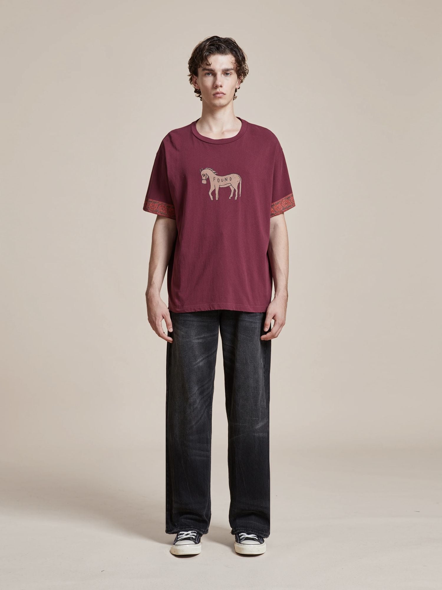 Person standing, wearing a dark red Found Horse Embellishment Tee with the word "HOUSE," black faded jeans, and black sneakers. The background is plain and light-colored.