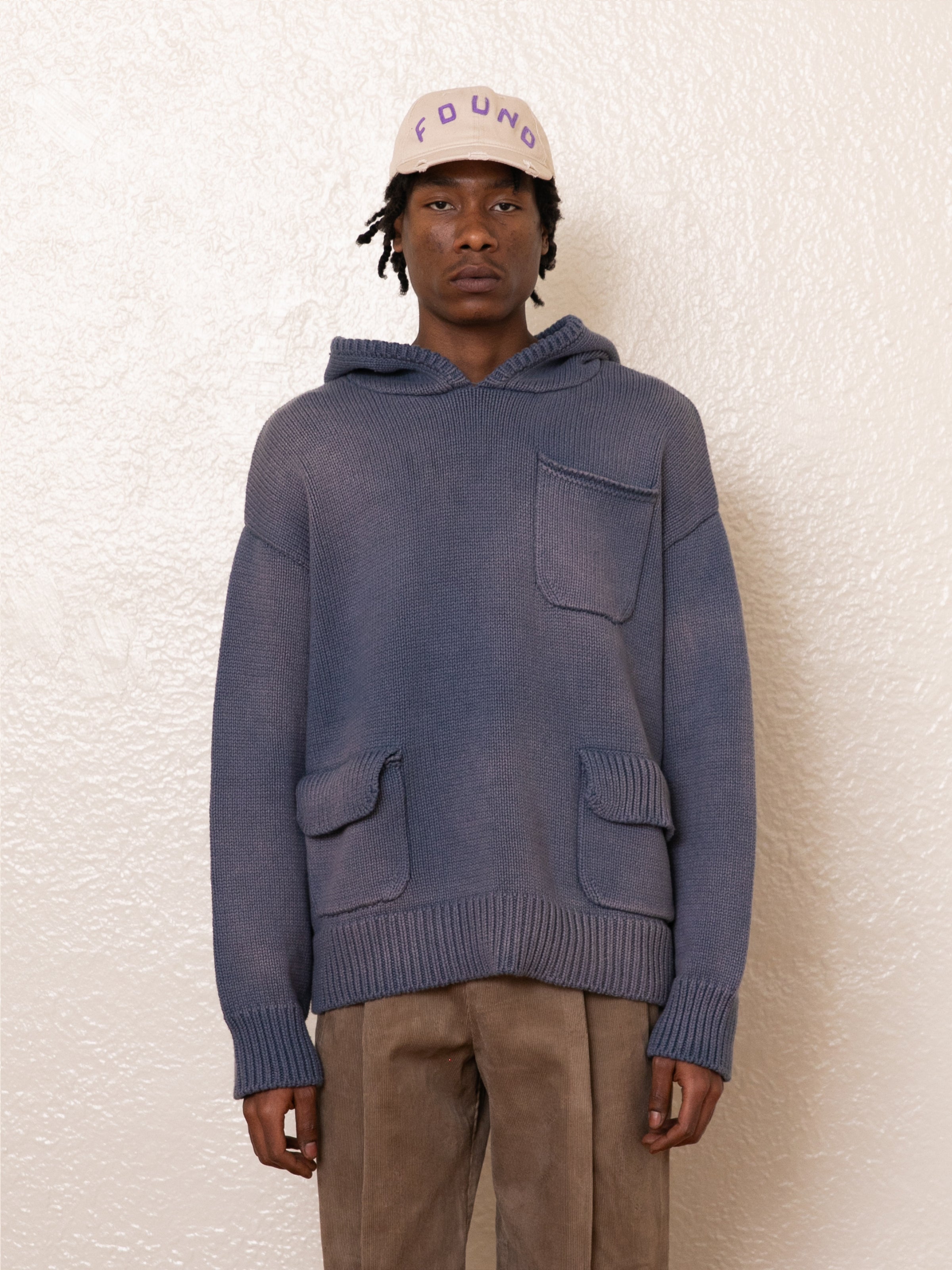 Against a textured white wall, an individual models FOUND's blue Faded Knit Pocket Hoodie with a relaxed oversized fit, paired with beige pants and accessorized with a light pink cap.