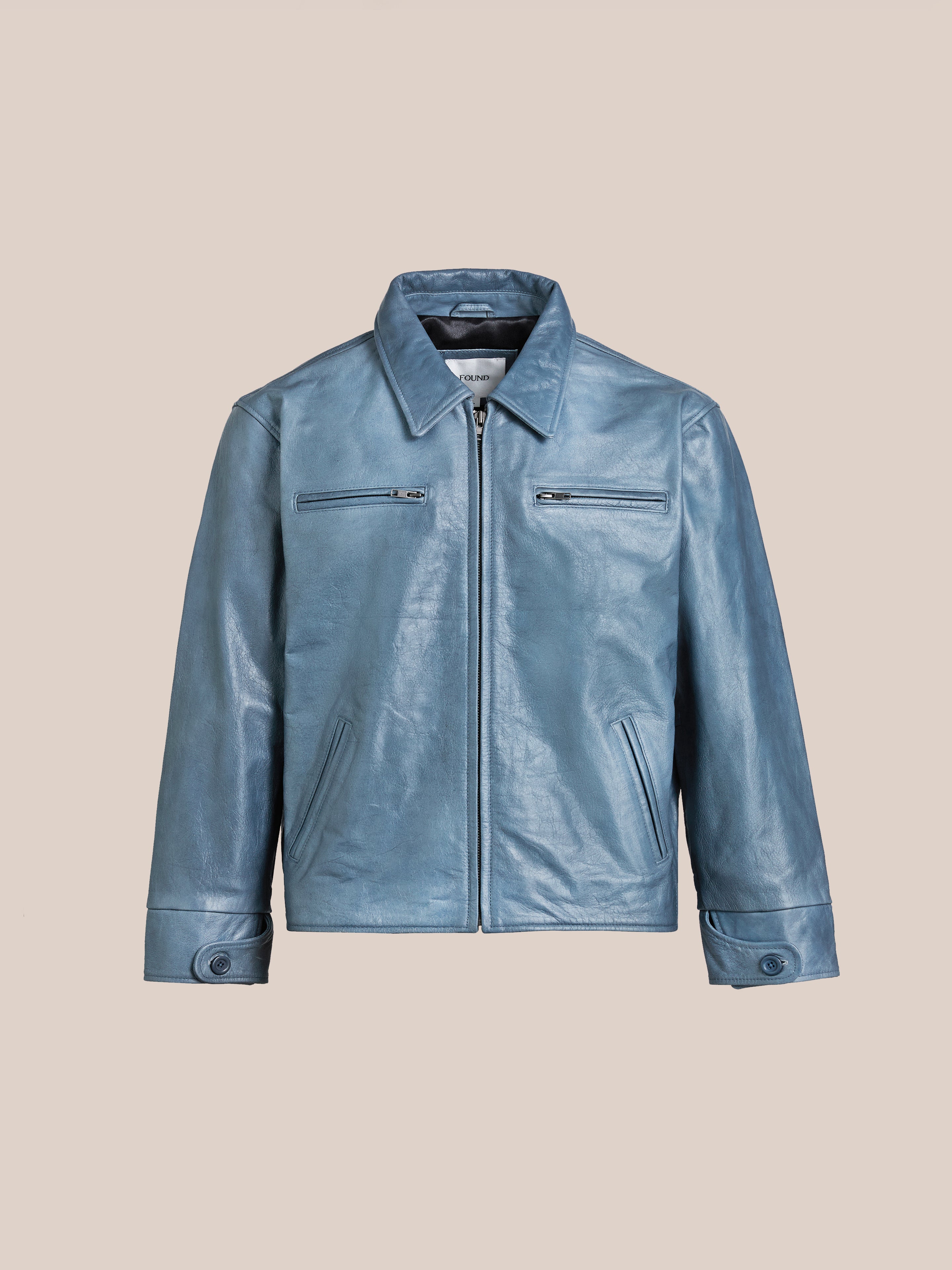 FOUND's Marlboro Sun Faded Leather Racer Jacket is a unisex light blue leather piece featuring a front zipper, fold-down collar, and two zippered chest pockets against a beige background.