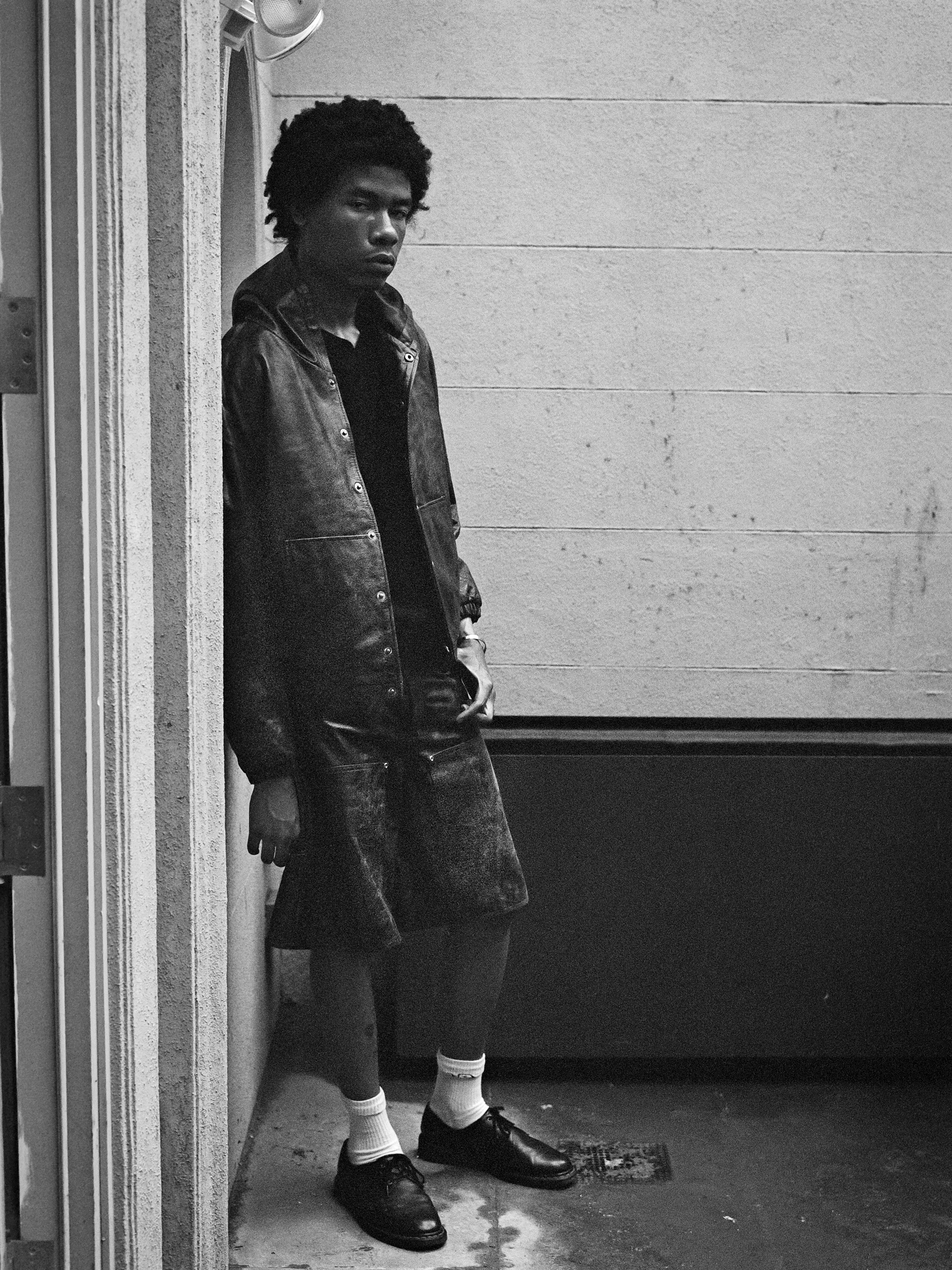 Black and white photo of a person standing against a wall, wearing a long leather coat, white socks, and black shoes. They have a serious expression and their hand in their pocket, showcasing the double-knee silhouette of their Distressed Genuine Leather Carpenter Shorts from Found, crafted from ethically sourced materials.