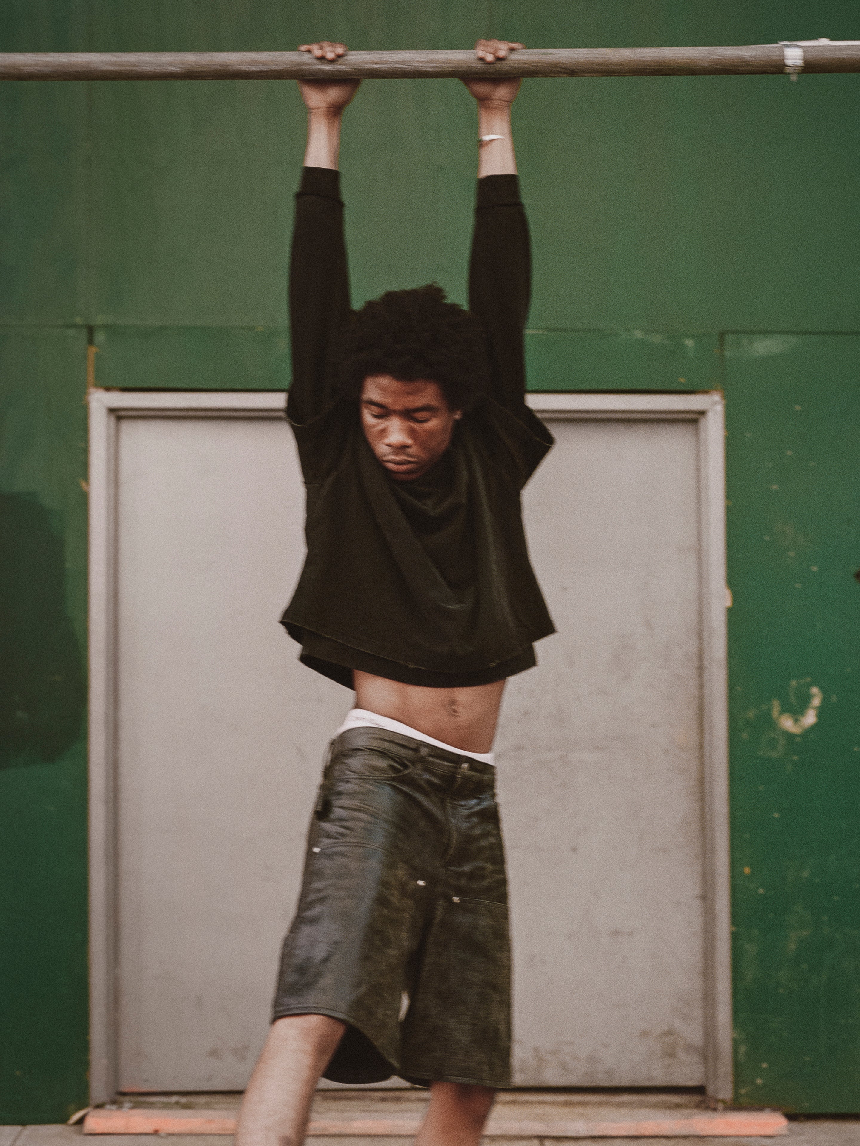 A person wearing a black long-sleeve shirt and Found's Distressed Genuine Leather Carpenter Shorts hangs from a horizontal bar in front of a green wall and a closed door.