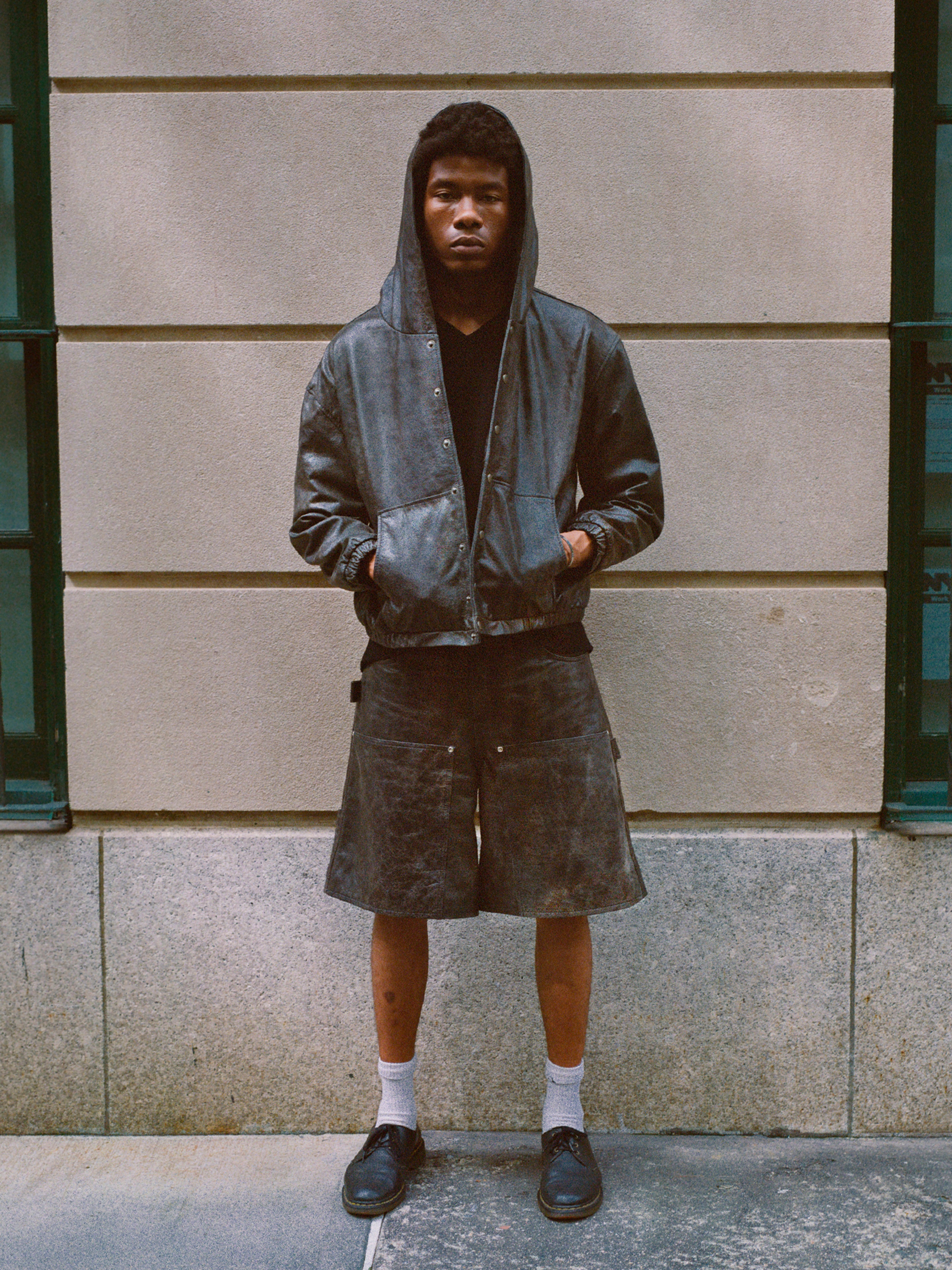 A person stands against a stone wall with hands in the pockets of a hooded jacket, wearing Found's Distressed Genuine Leather Carpenter Shorts featuring a double-knee silhouette, white socks, and black shoes.