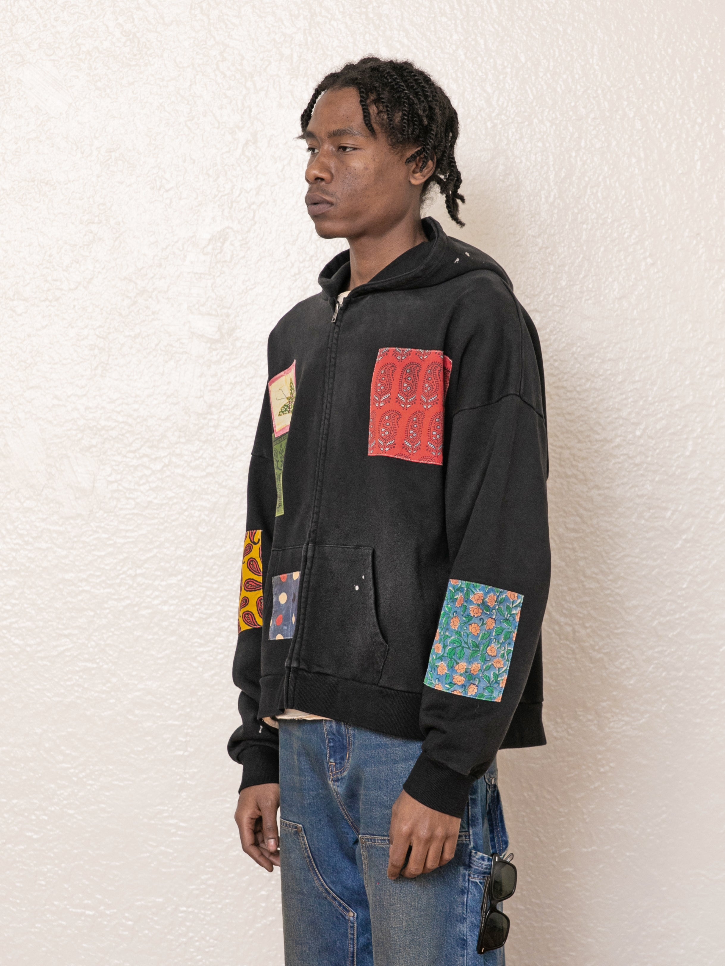 A person wearing FOUND's Tapestry Patchwork Zip Up Hoodie, featuring colorful vintage-inspired designs, stands against a textured white wall to showcase its oversized fit.