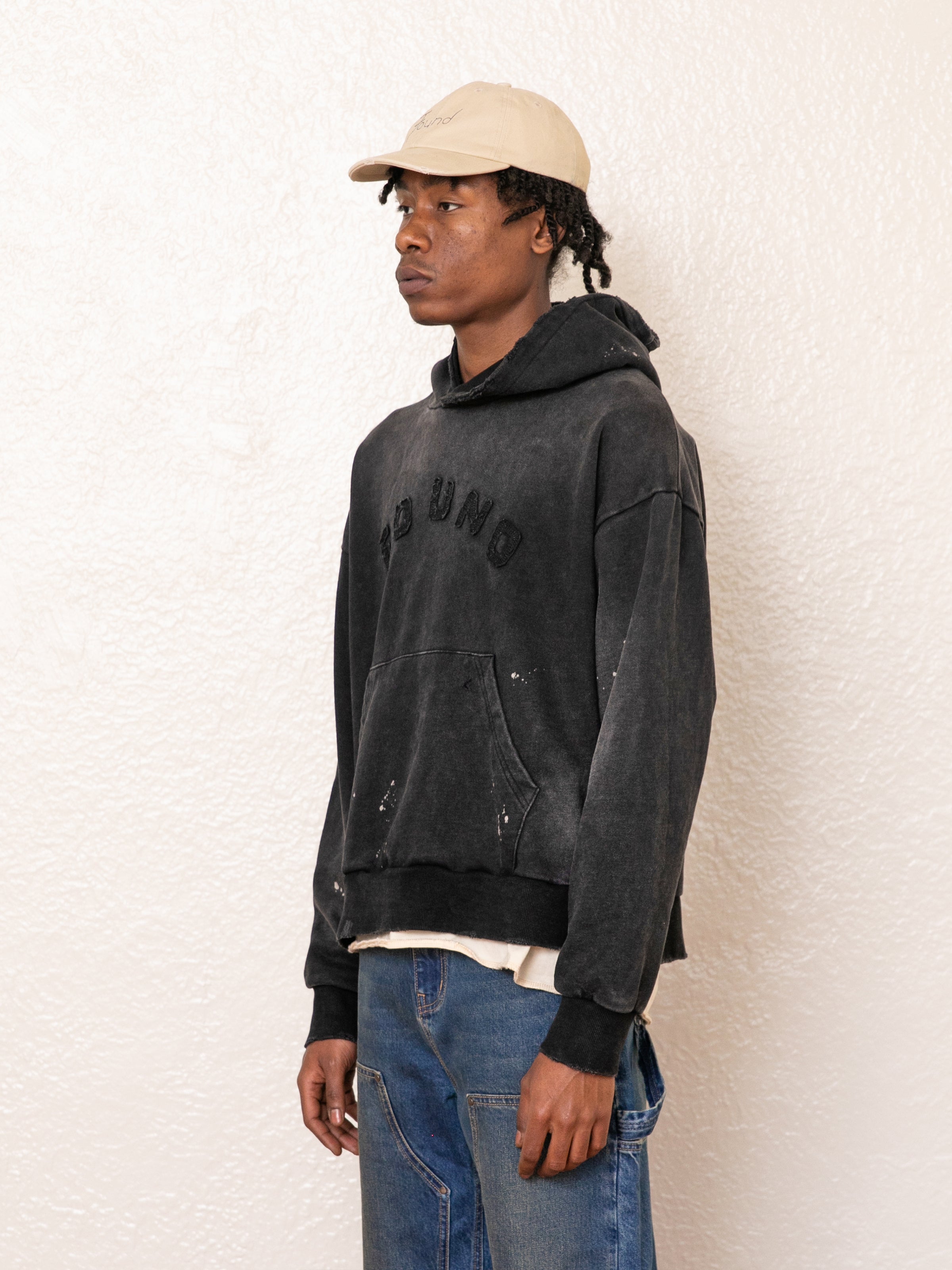 A person wearing a FOUND beige cap and vintage black Logo Patch Hoodie made from French terry cotton stands against a textured white wall, looking left and effortlessly paired with blue jeans.