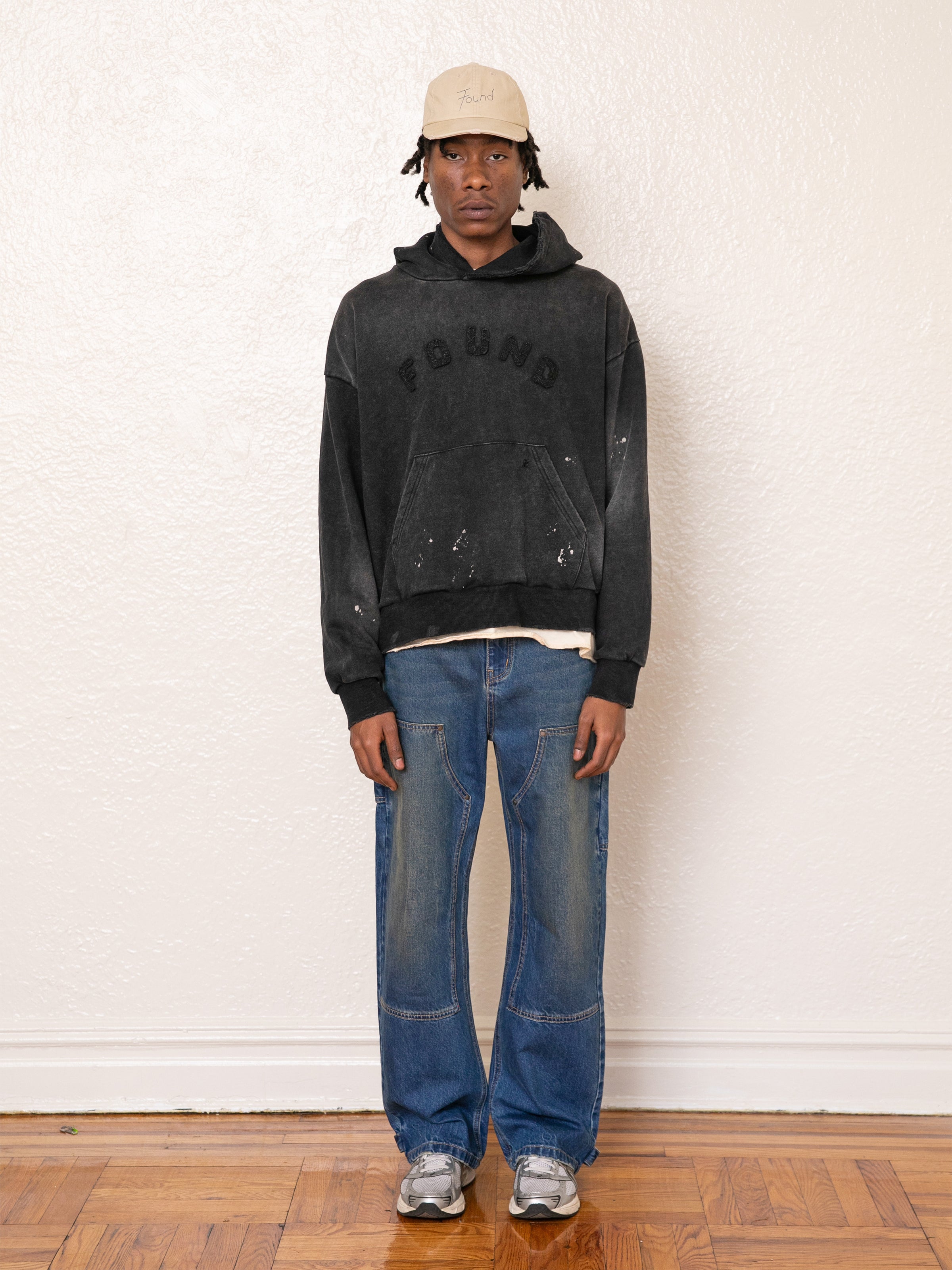 A person in a vintage black FOUND Logo Patch Hoodie, blue jeans, sneakers, and a beige cap stands against a plain wall on a wooden floor.